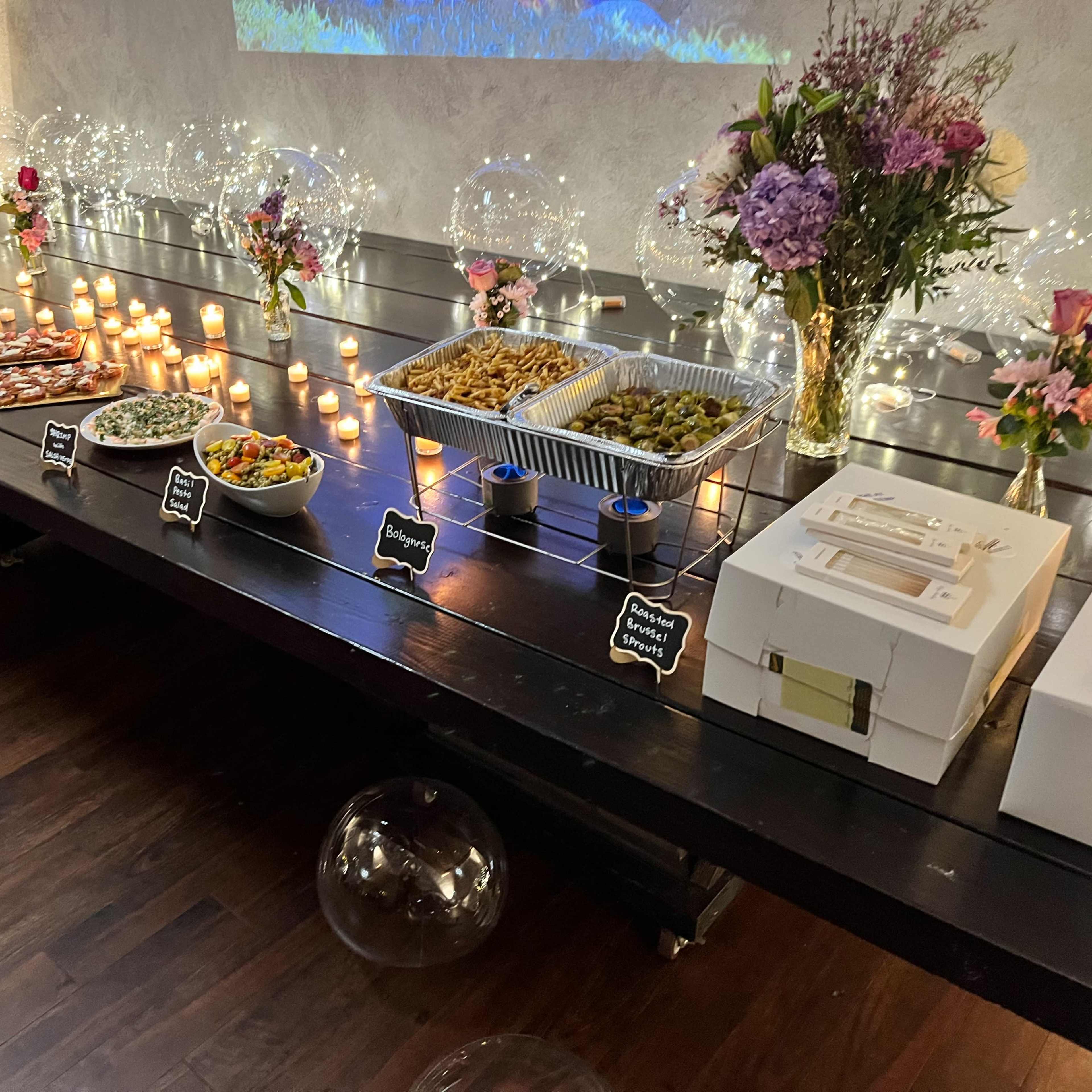 A long wooden table displays an array of food items, candles, and floral arrangements against a softly lit background.