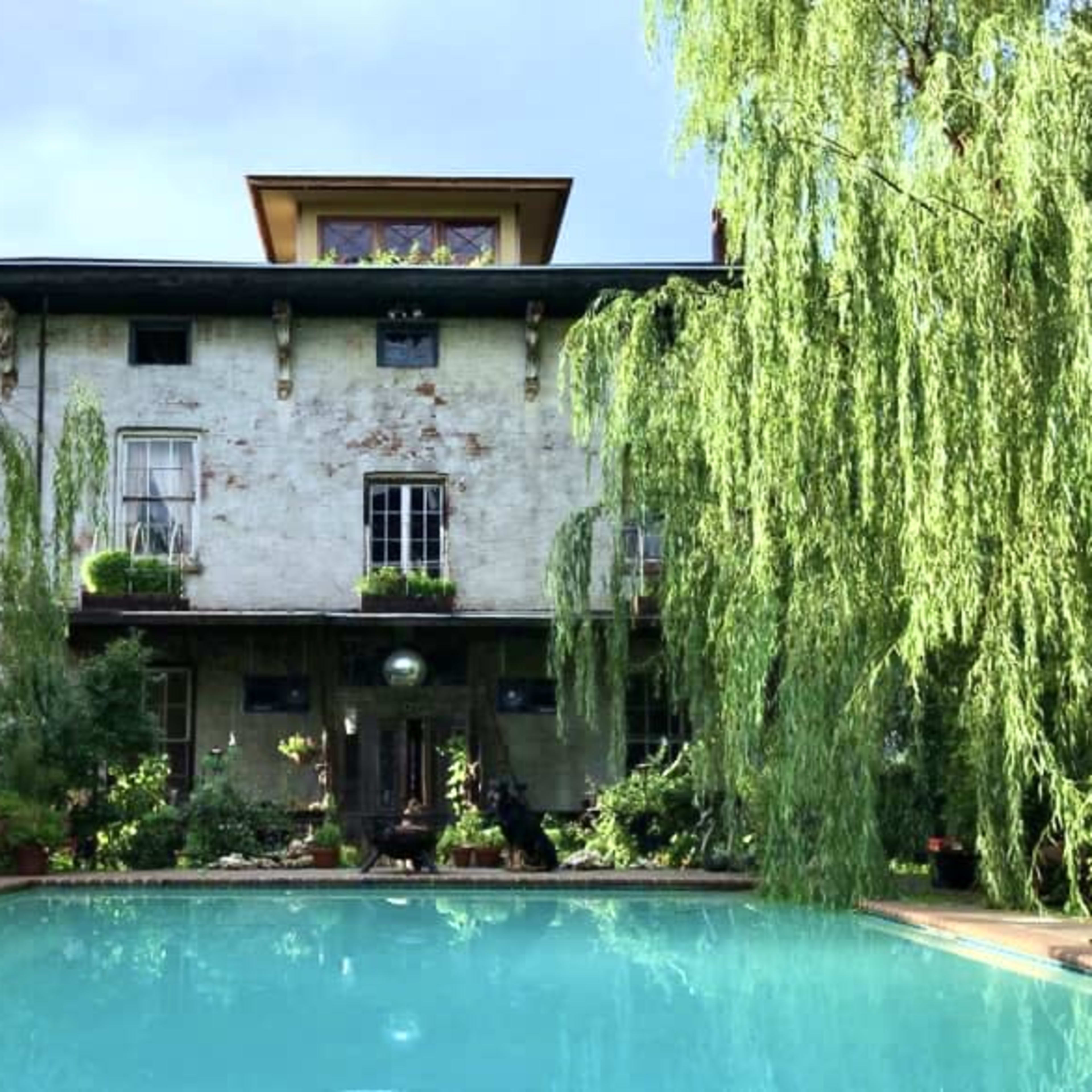 A historic brick building with tall windows is situated behind a turquoise swimming pool and surrounded by weeping willow trees.