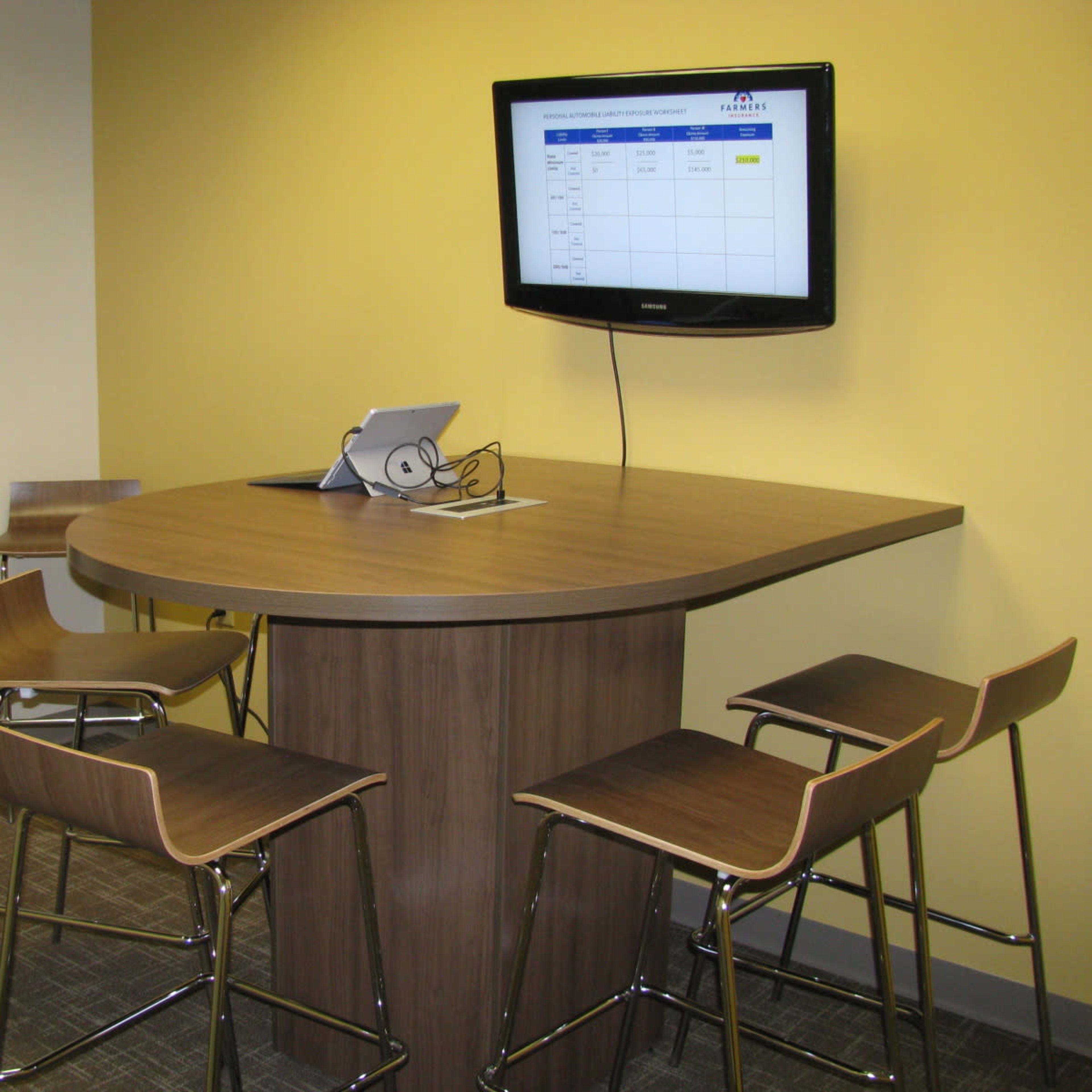 A modern conference area features a brown table with high stools and a wall-mounted television displaying a spreadsheet.