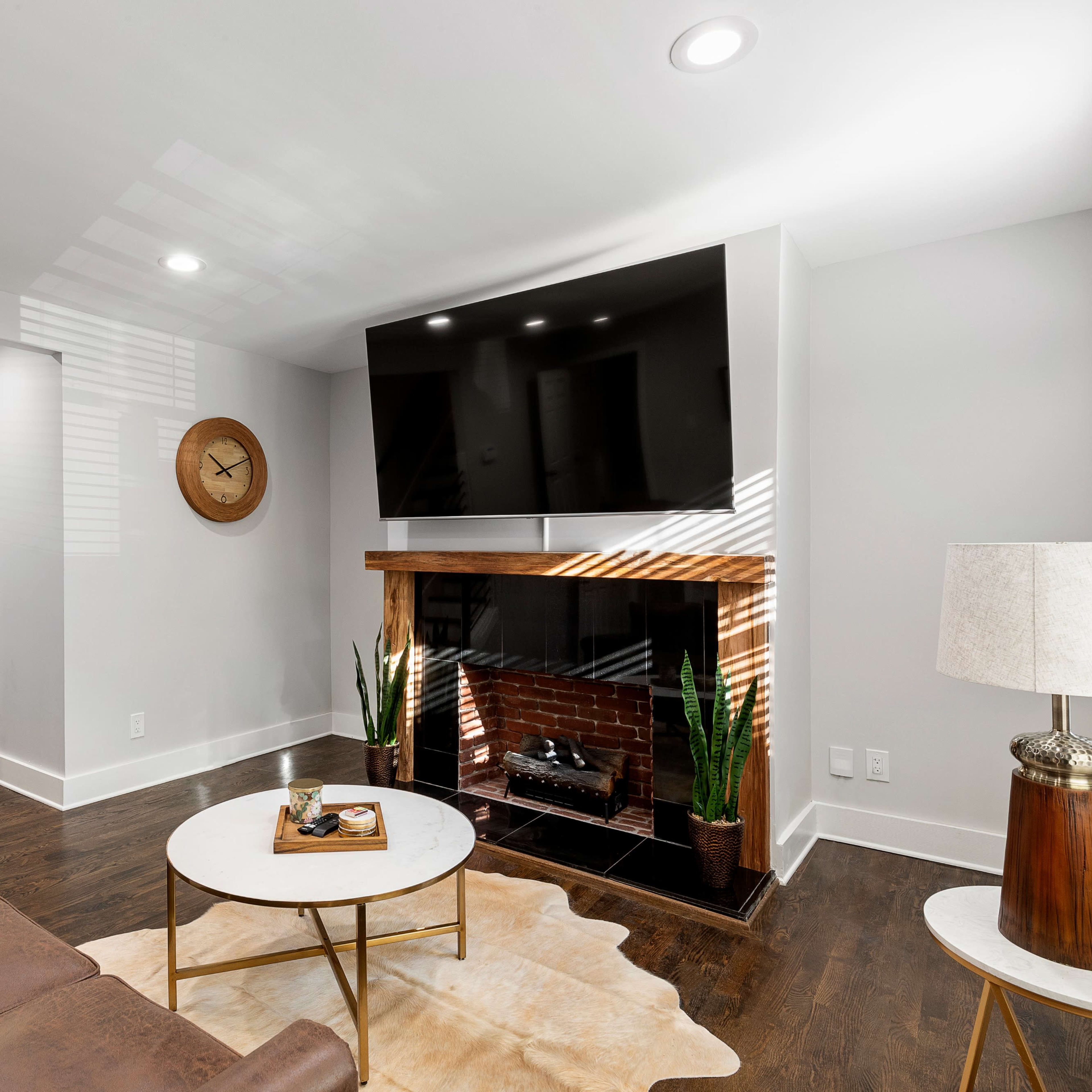 A modern living room with a brown leather sofa, a round coffee table, a wall-mounted television above a brick fireplace, and plants in the corner.