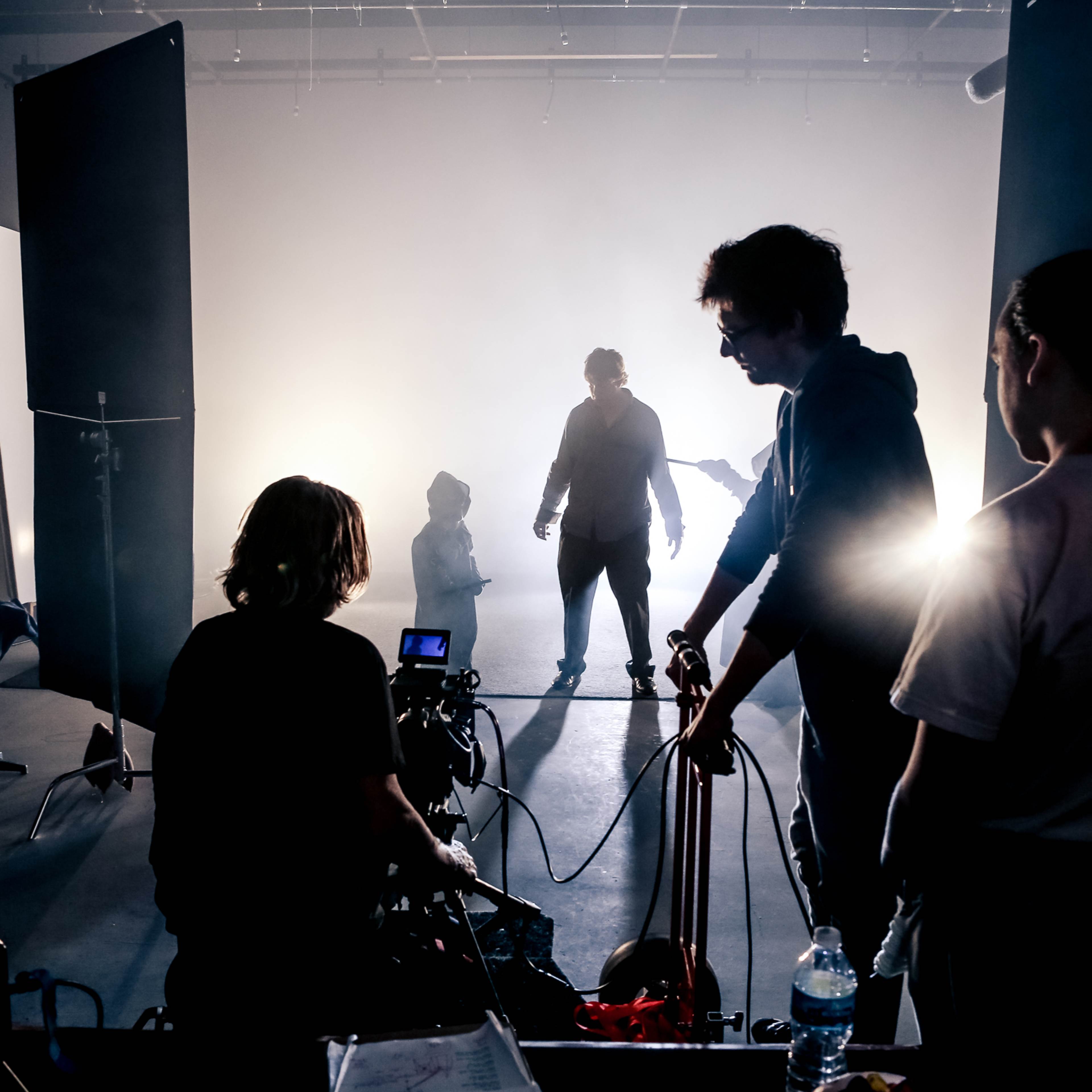 A film crew works in a dimly lit studio, with silhouetted figures and equipment set up for a shoot.