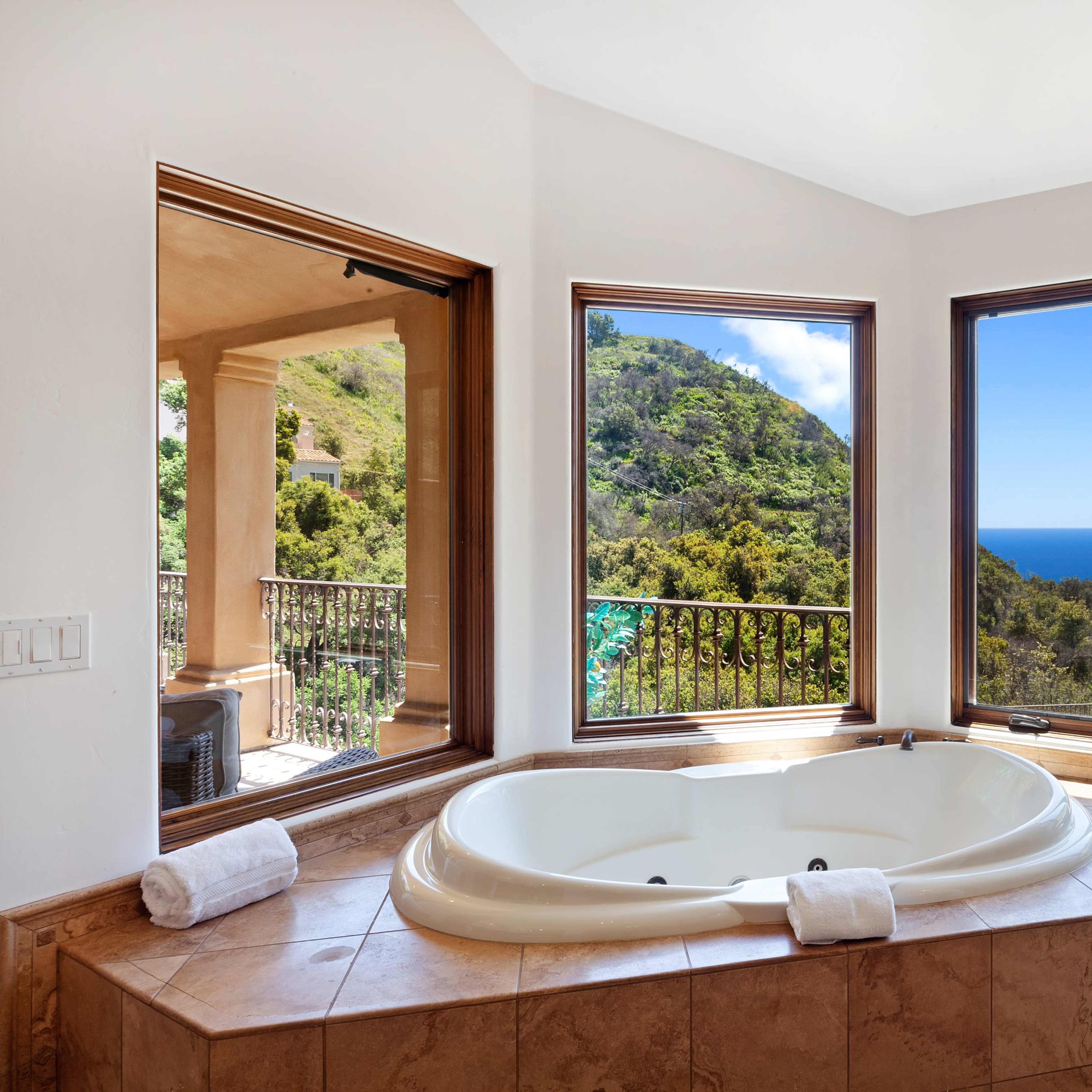 A spacious bathroom features a sunken jacuzzi surrounded by large windows offering a view of lush hills and the ocean.
