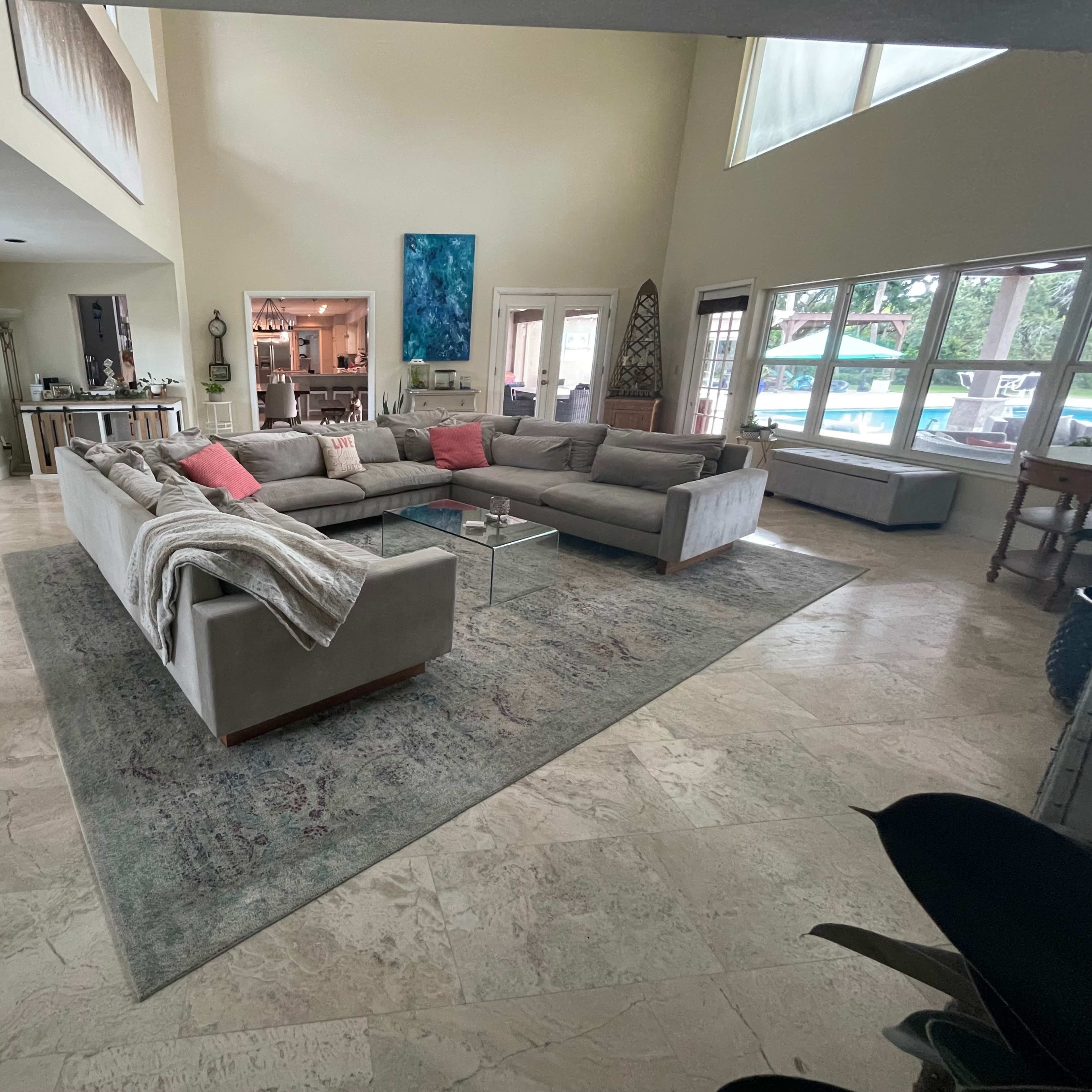 A spacious living room features a large sectional sofa arranged around a glass coffee table, with large windows allowing natural light and a view of an outdoor area.