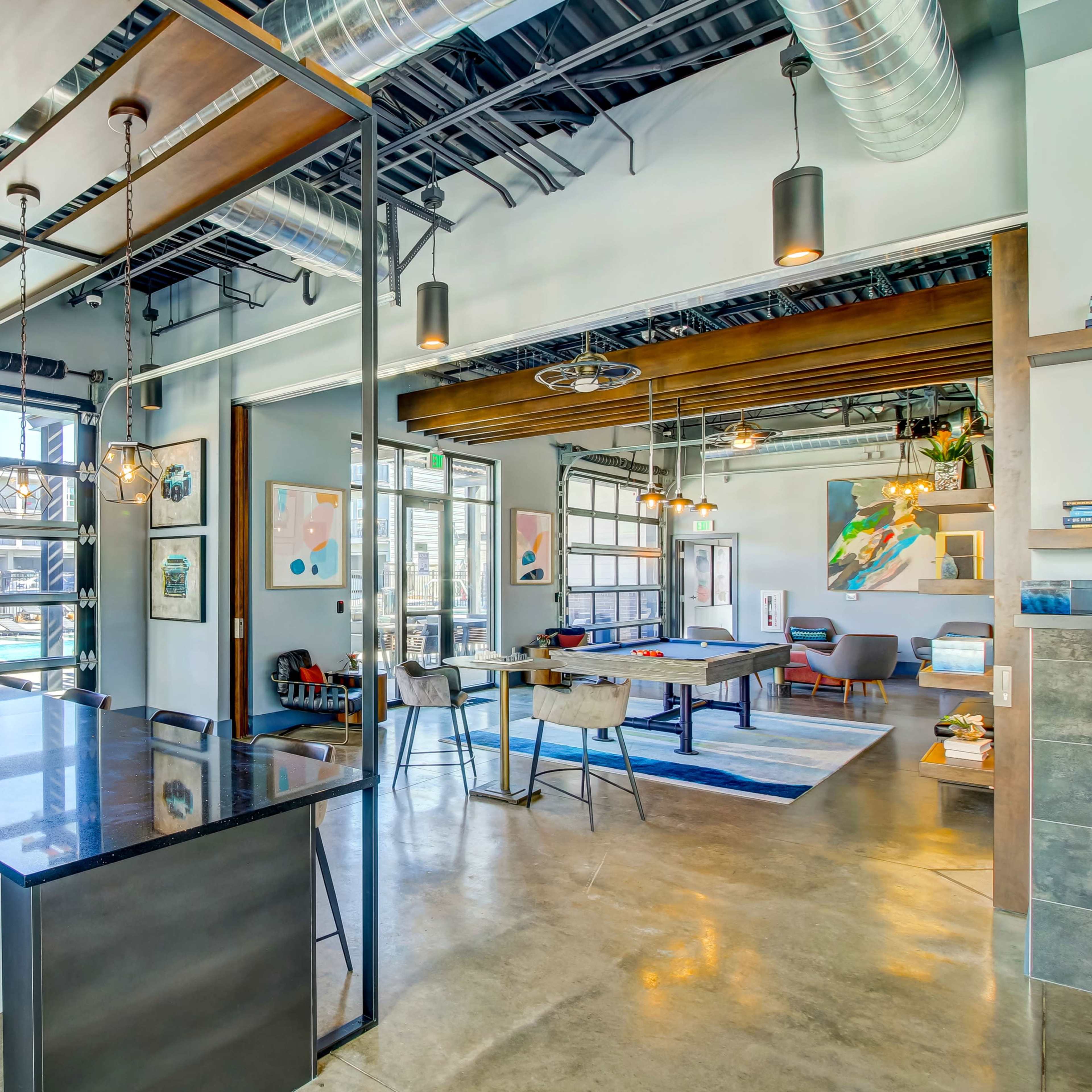 A modern open-concept office space with large windows, industrial lighting, and a kitchen area adjacent to collaborative workspaces.