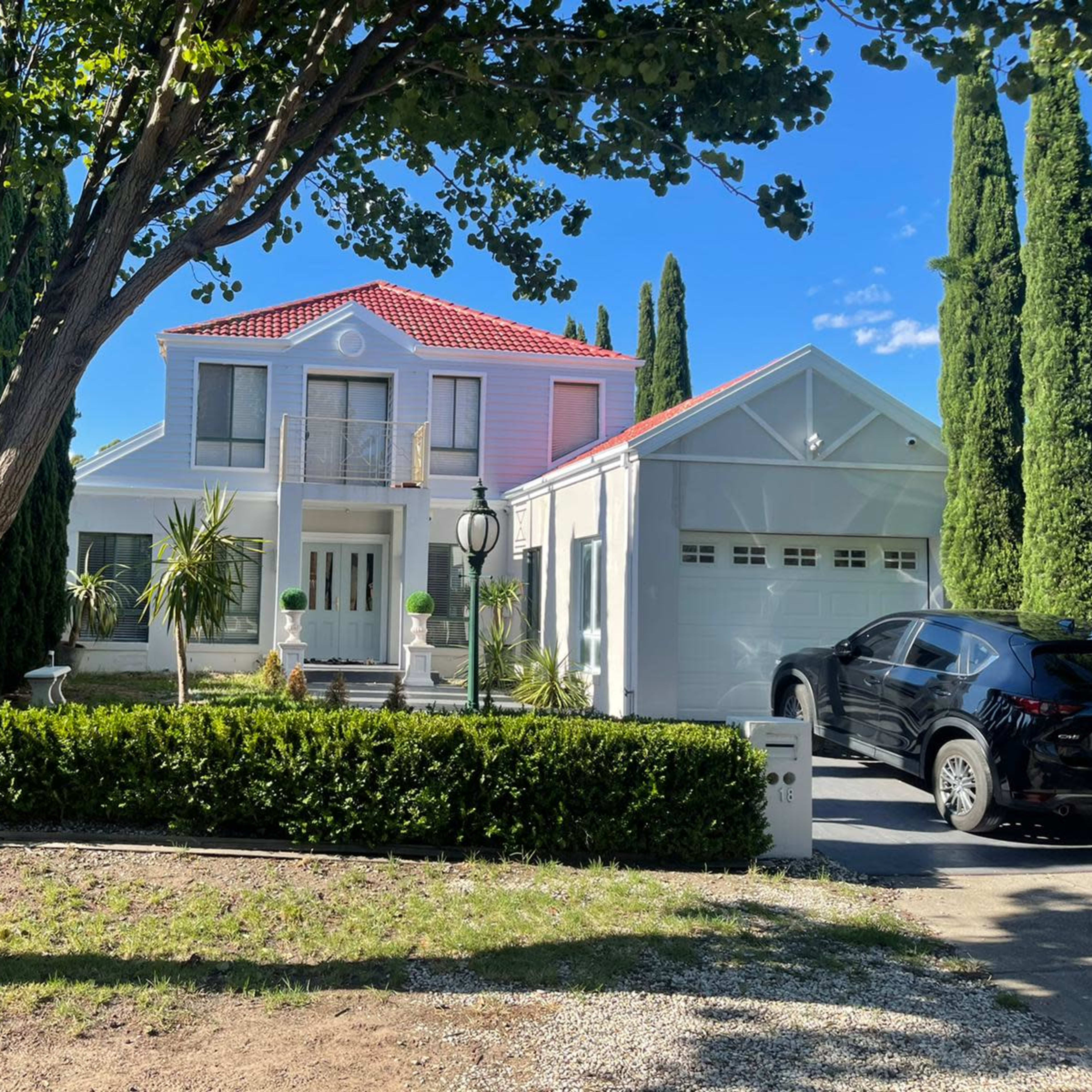 A two-story house with a red-tiled roof is surrounded by tall trees and shrubs, with a garage on the right and a car parked in the driveway.