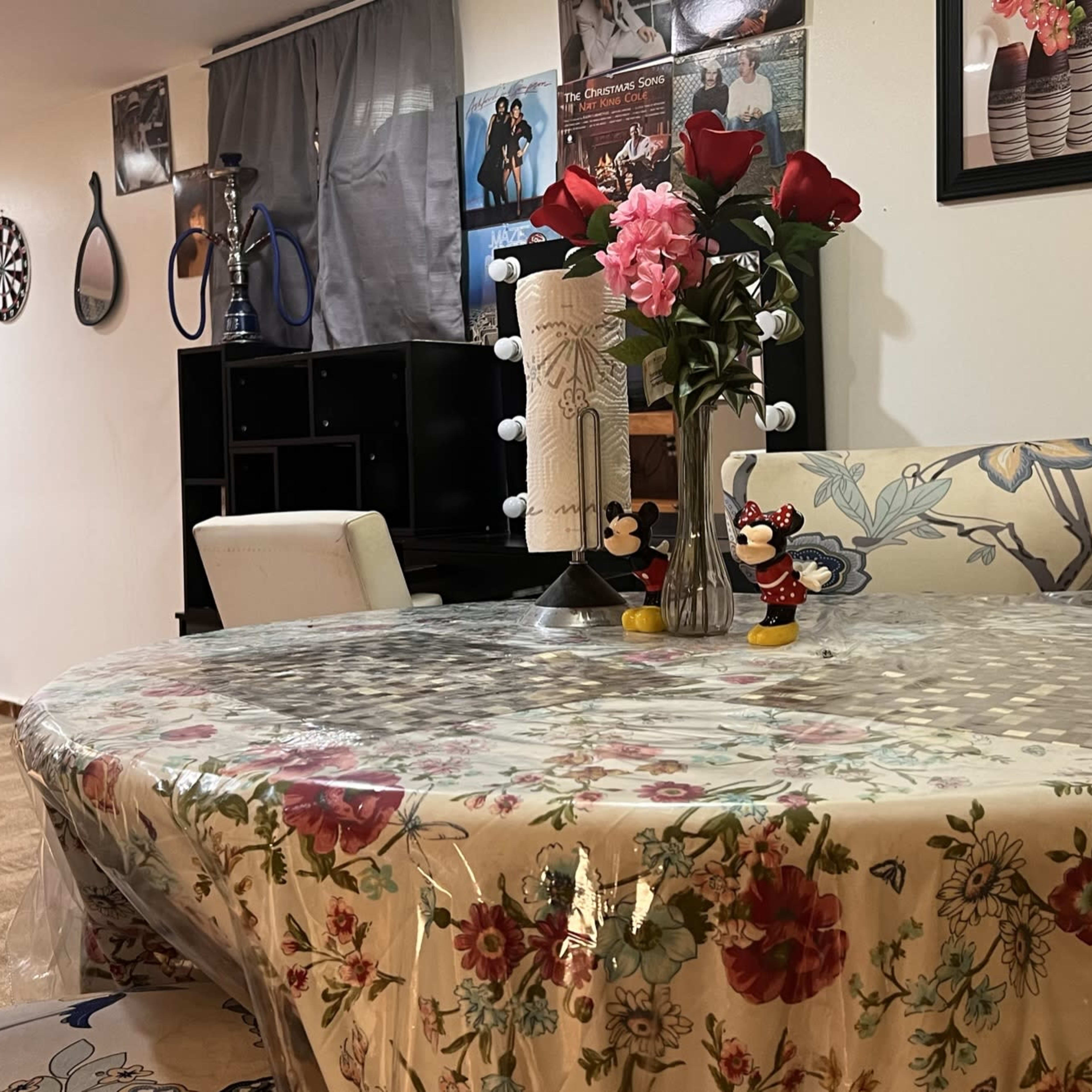 A cozy, semi-furnished room with a dining table set with a floral tablecloth, a vase of roses, and decorative items, alongside a bed in the background and posters on the walls.