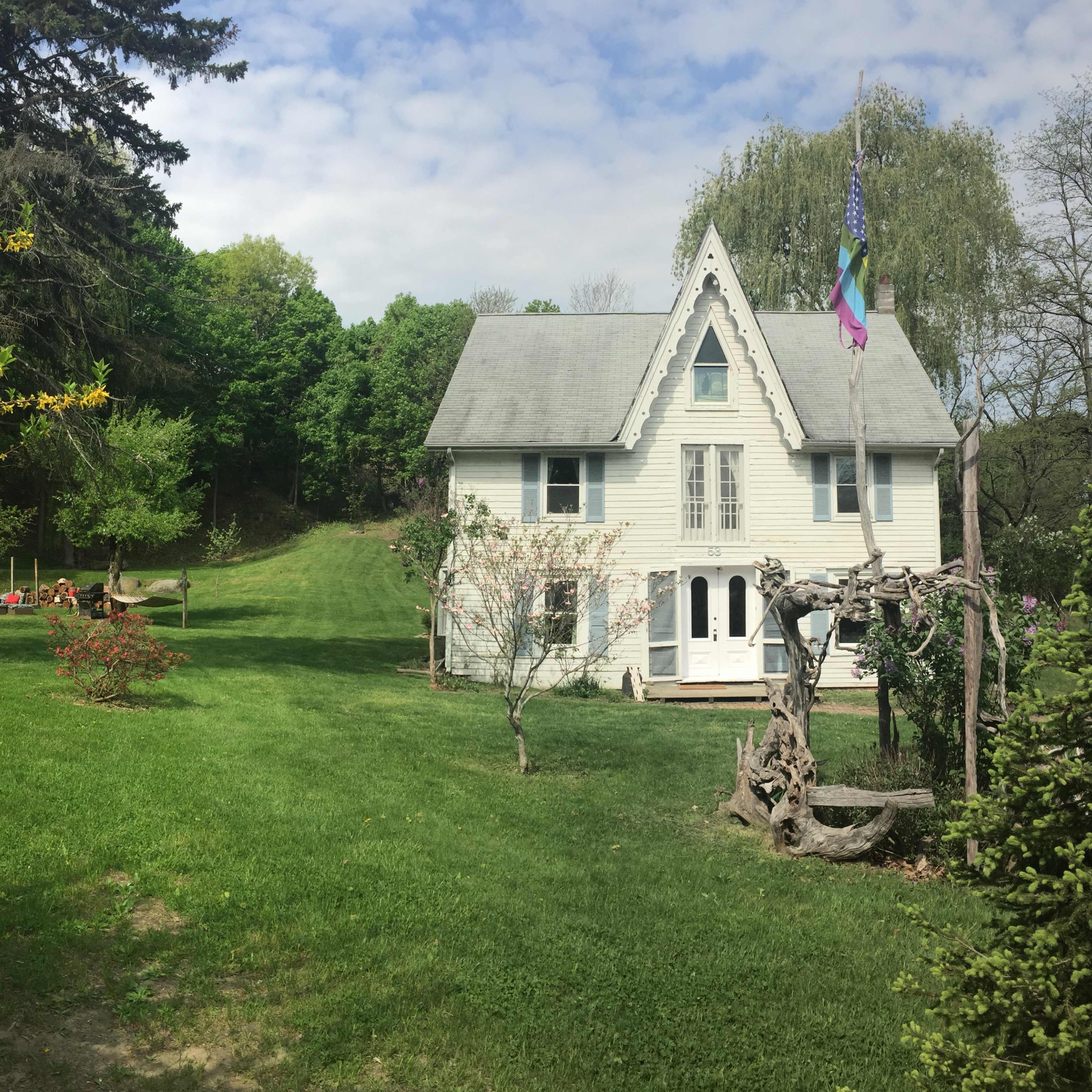 A two-story house with a pointed roof and flags stands in a green yard surrounded by trees and a wooden swing set.