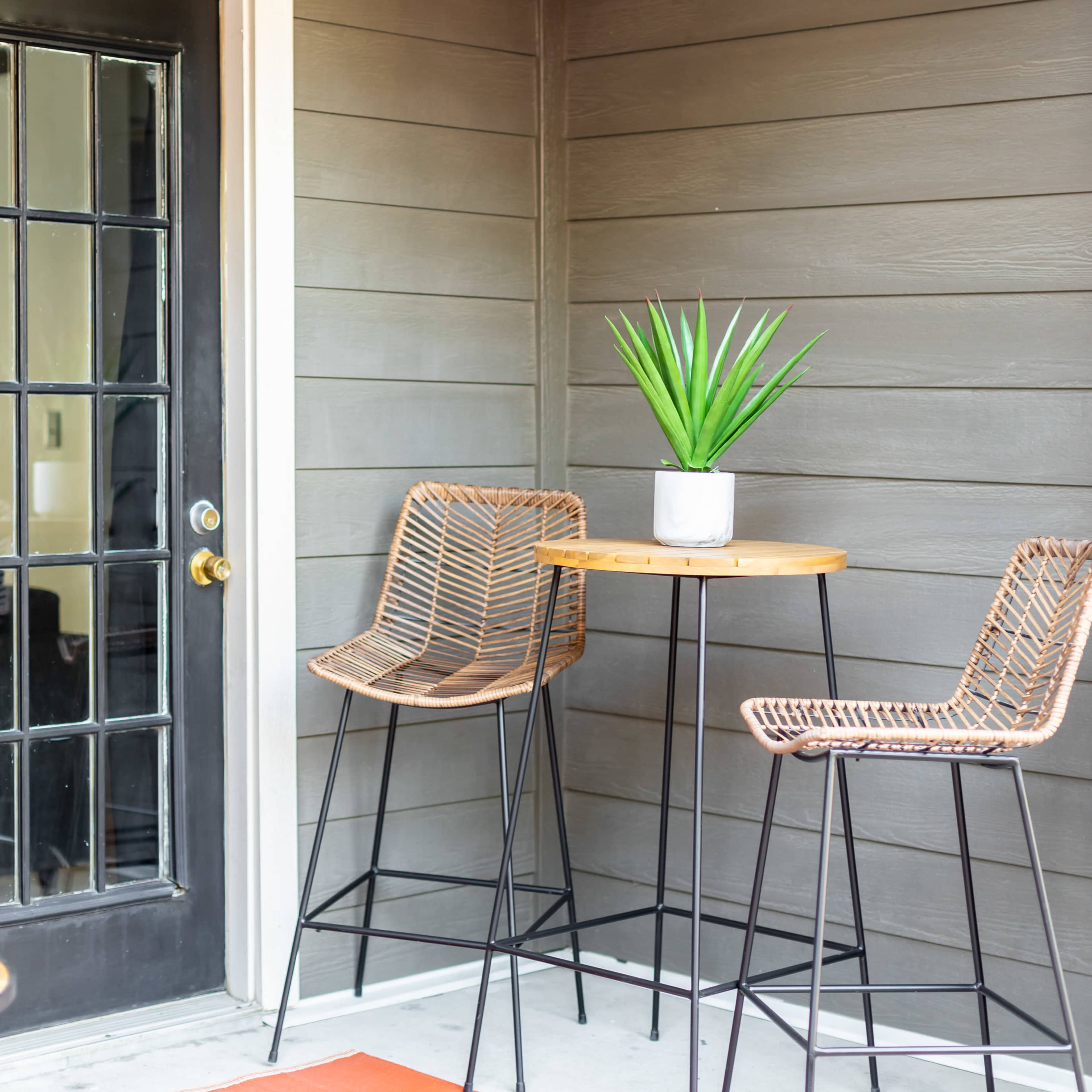A small outdoor seating area features two high stools and a table with a potted plant on it, beside a door with glass panels.