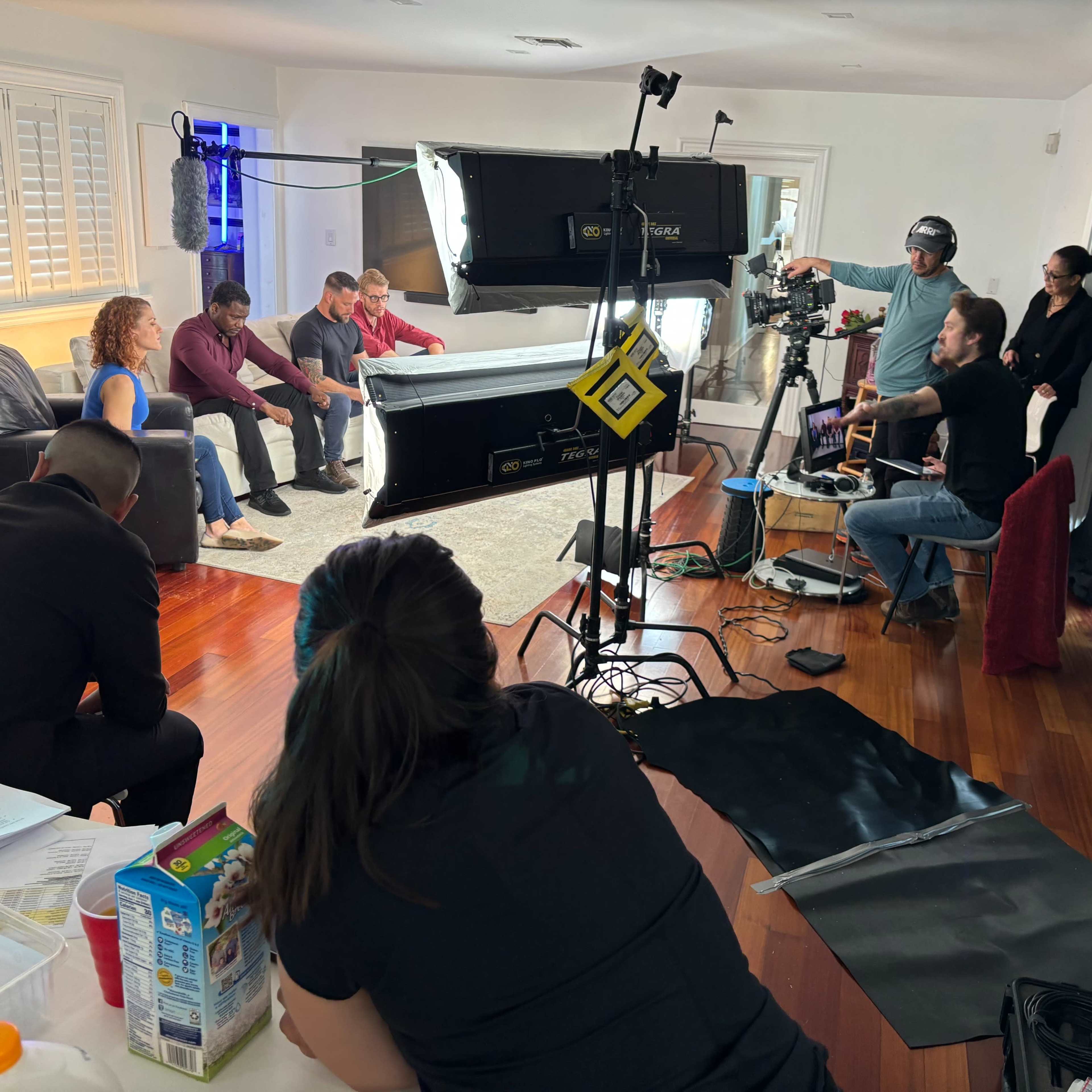 A film crew sets up equipment and prepares for a shoot in a well-lit living room.