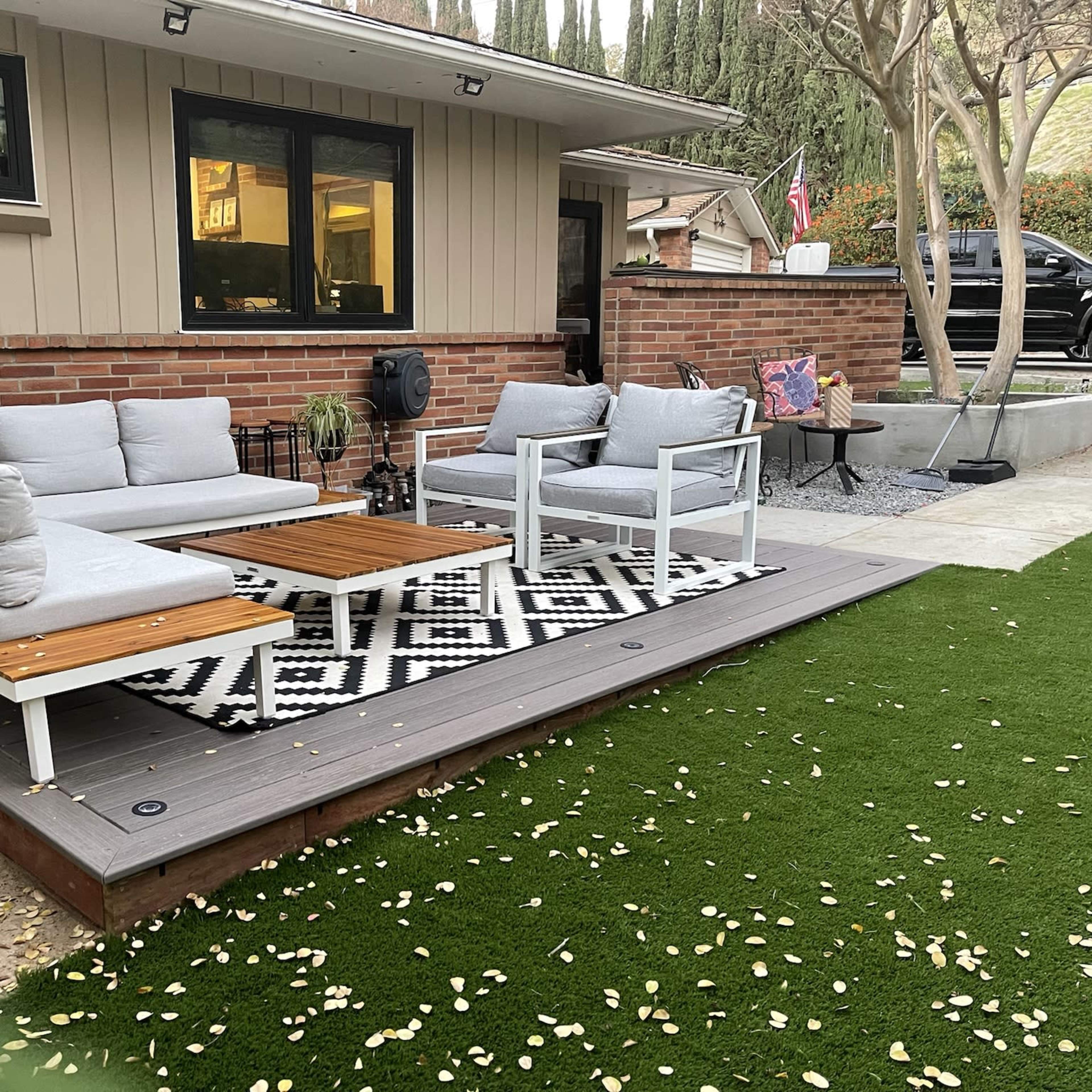 A outdoor seating area features a wooden deck with gray couches and a patterned rug surrounded by grass and landscaping.
