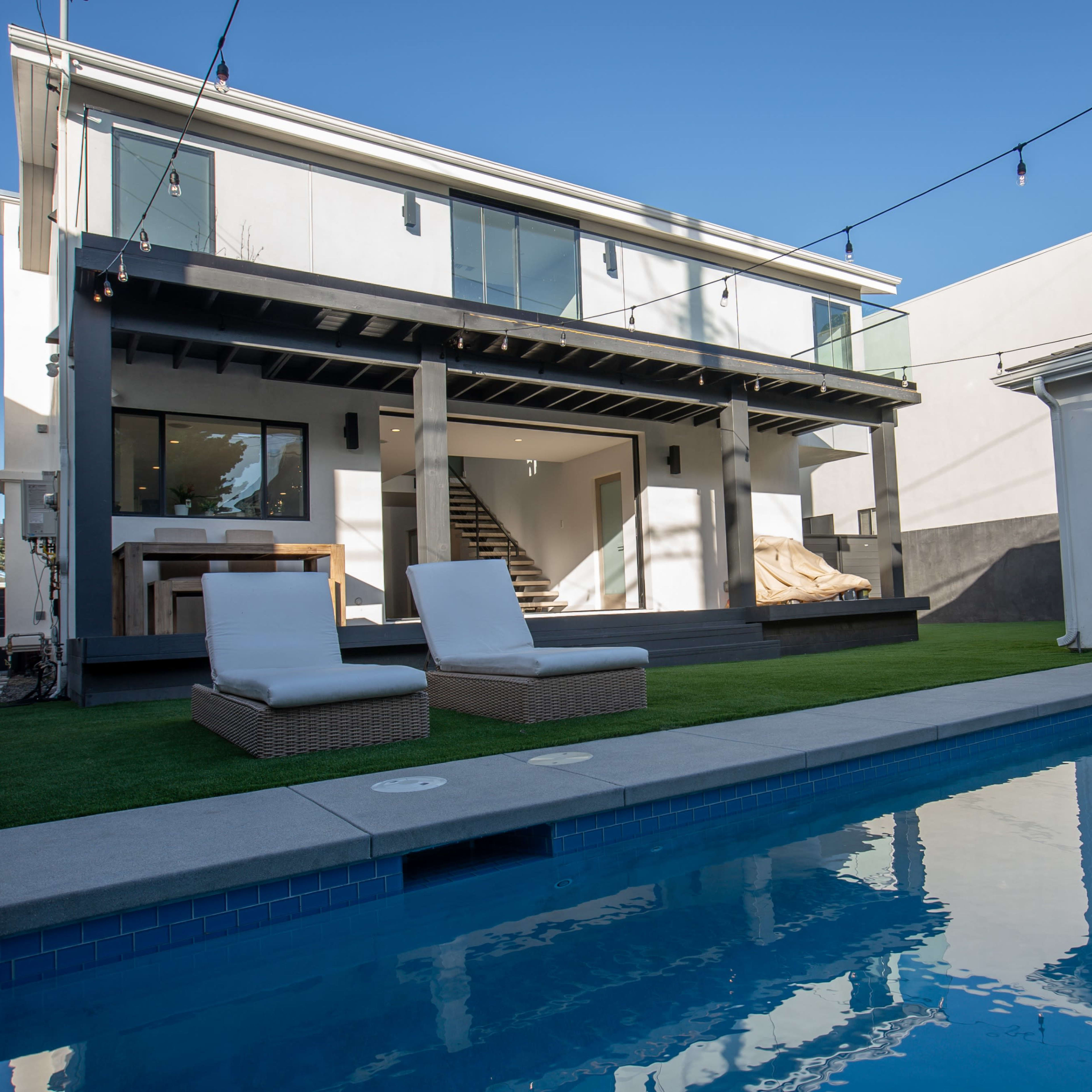 A modern two-story house with a pool, outdoor seating, and stairs leading to a covered patio is set against a clear blue sky.