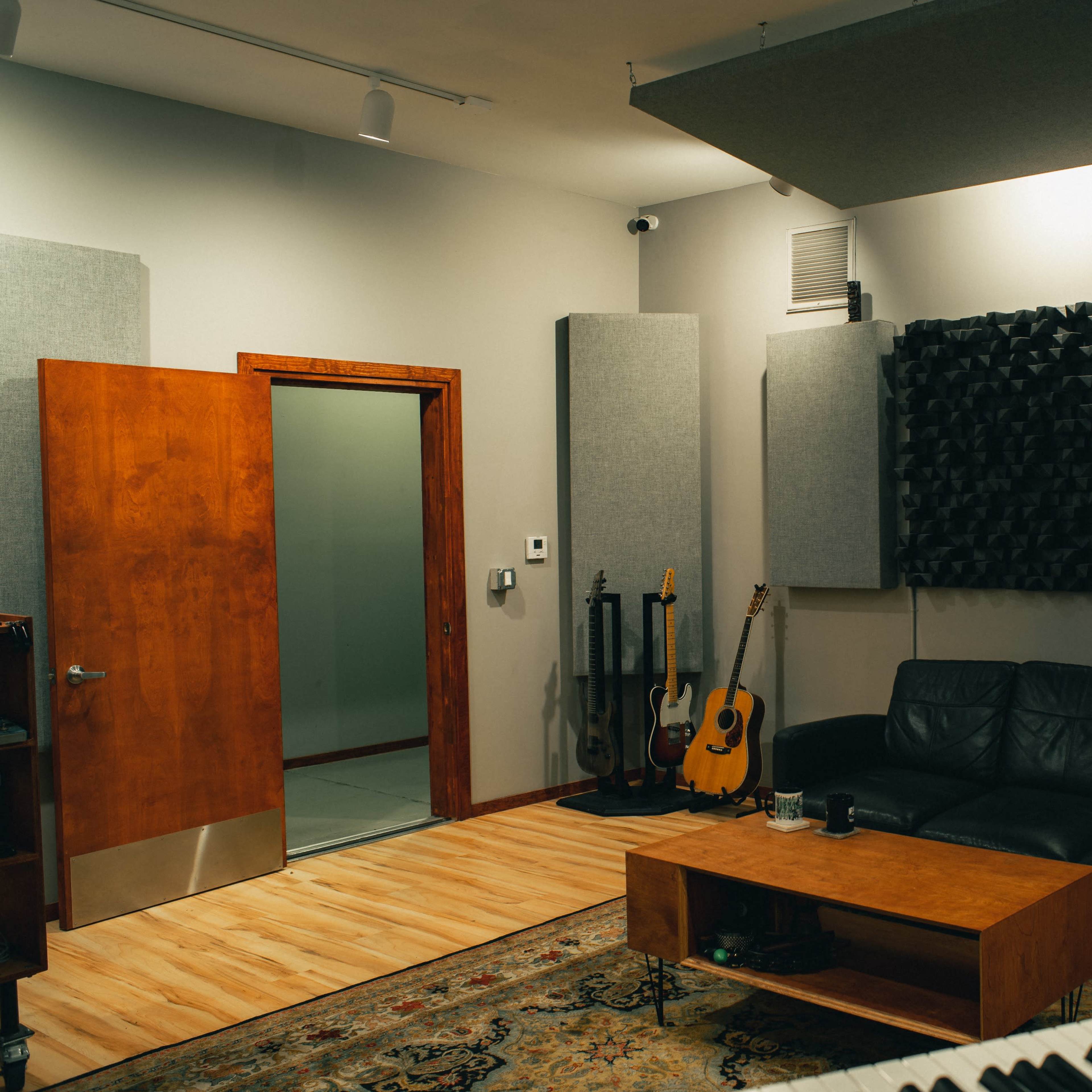 A music studio featuring a black sofa, a wooden coffee table, acoustic panels on the walls, and various musical instruments leaning against the wall.