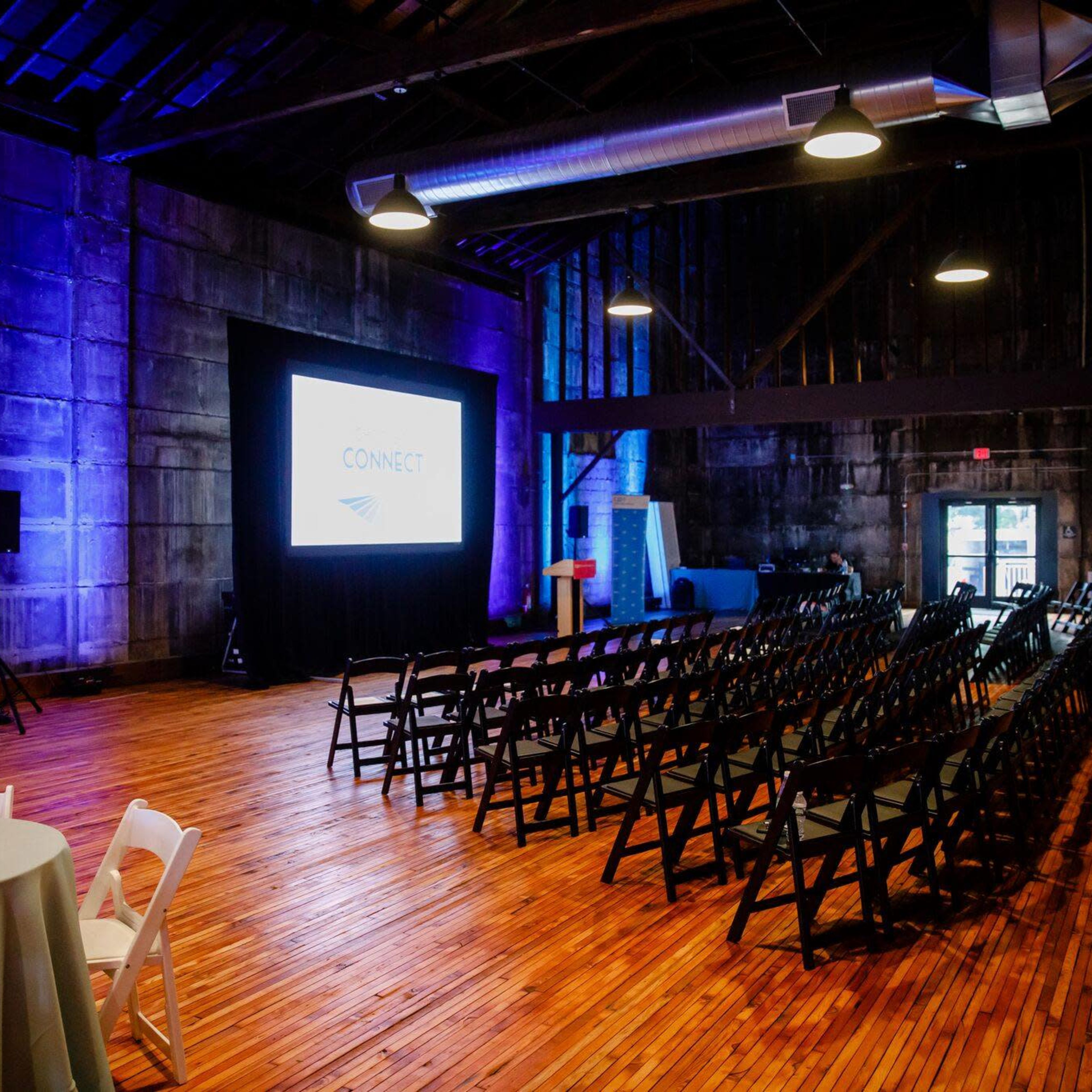 A dimly lit event space with wooden flooring, featuring rows of black chairs facing a screen, surrounded by round tables.