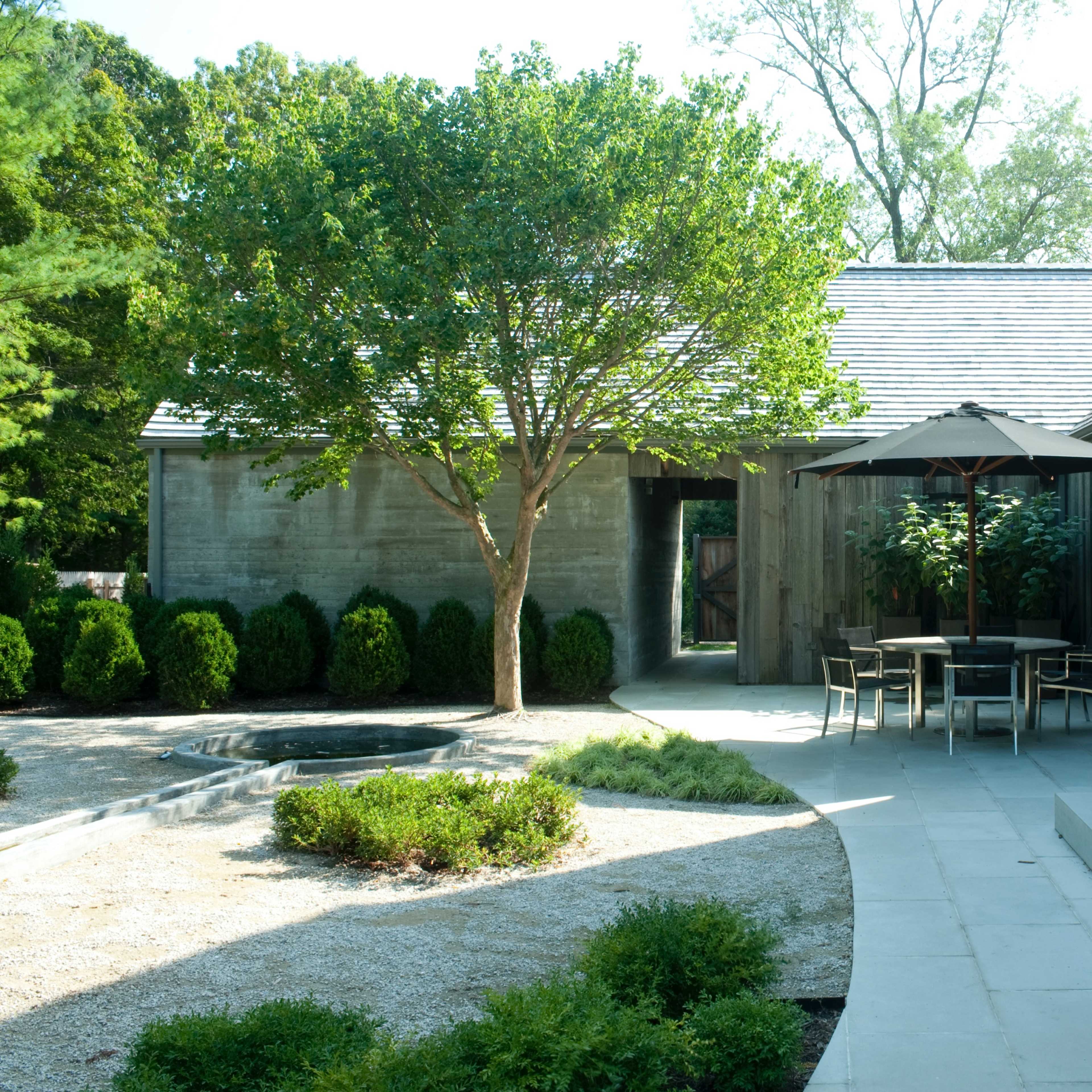 A modern outdoor patio area with a dining table and an umbrella, surrounded by greenery and a pathway leading to a house with large open doors.
