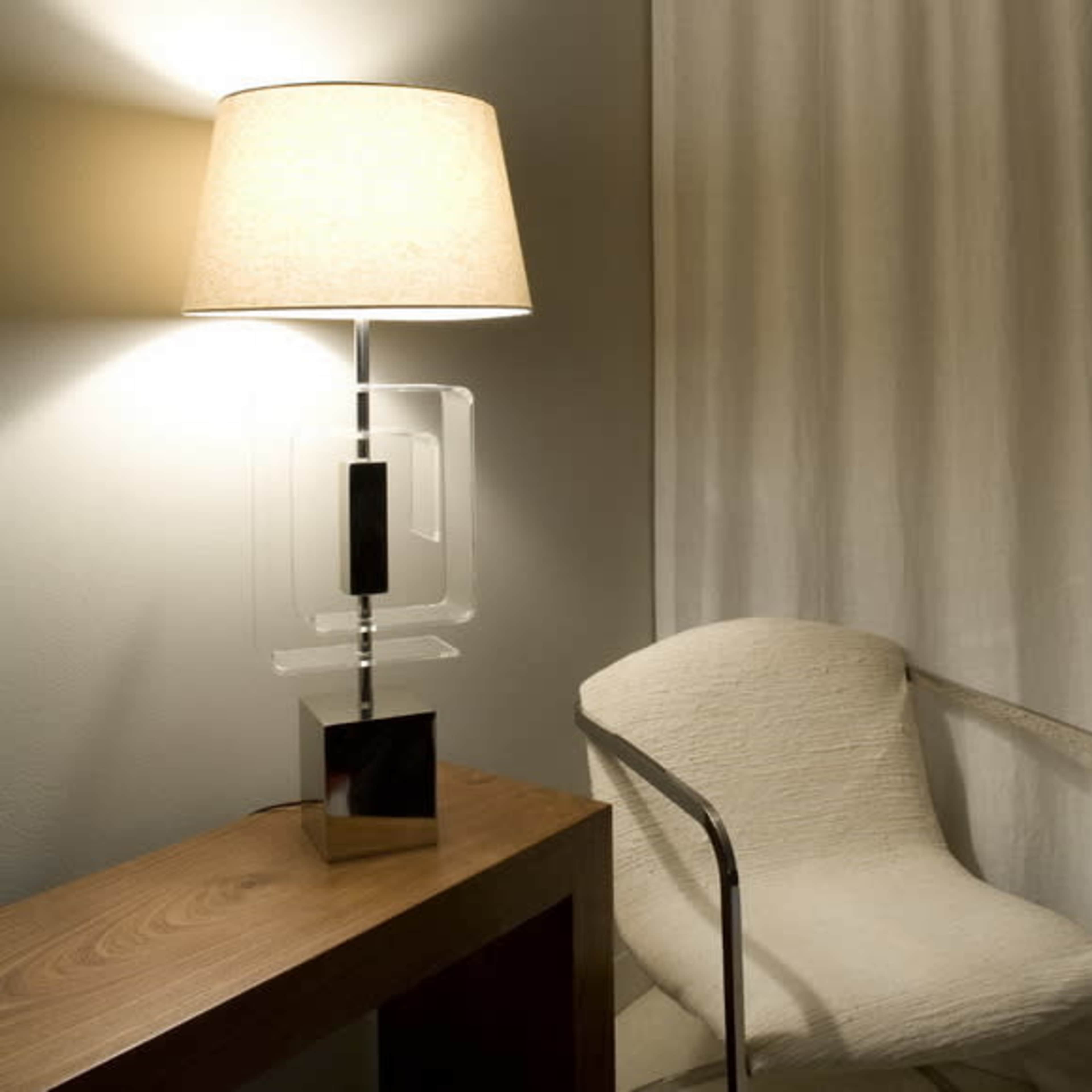 A modern lamp with a beige shade stands on a wooden table next to a white chair in a softly lit room with white curtains.