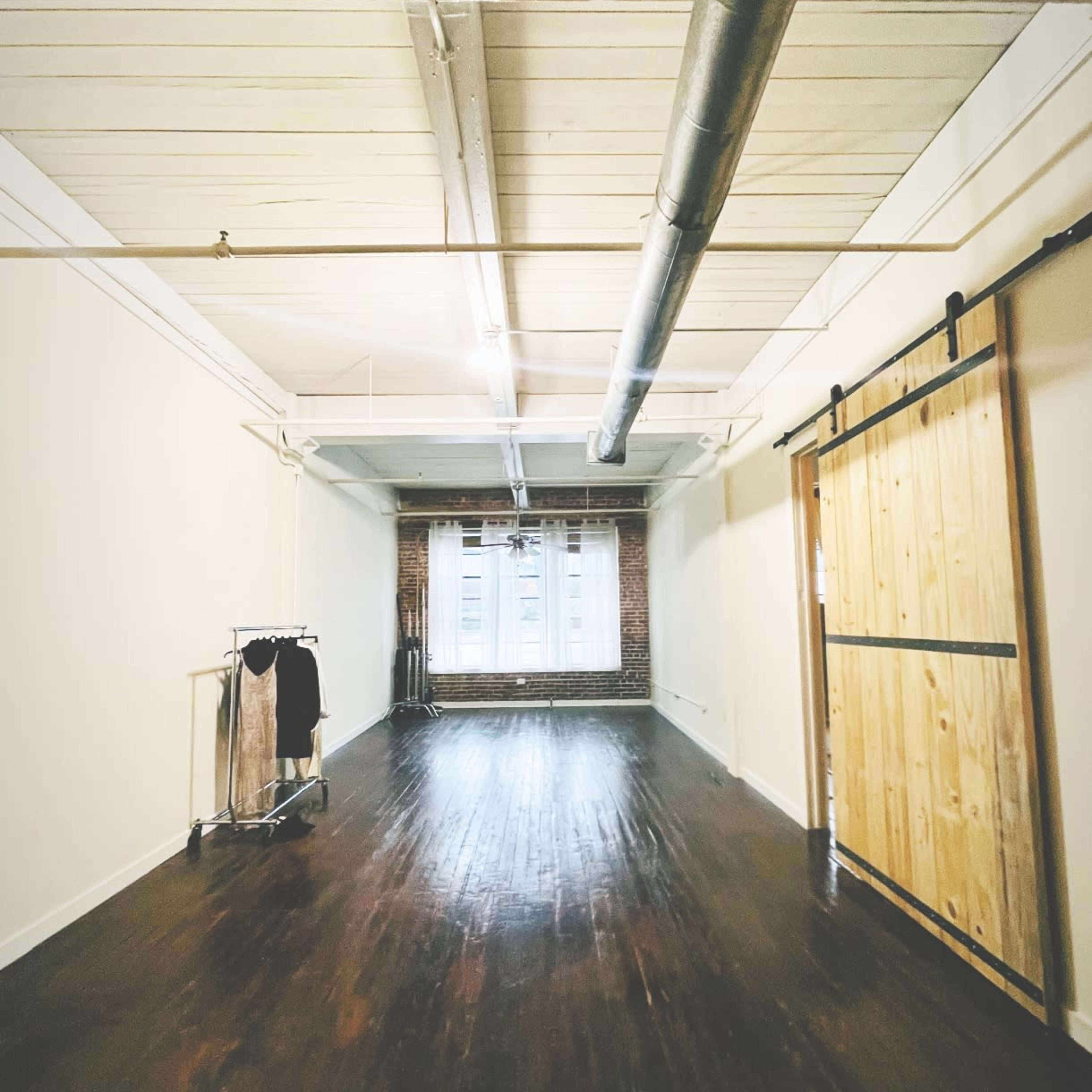 A spacious room features wooden floors, exposed brick walls, and a large window with sheer curtains.