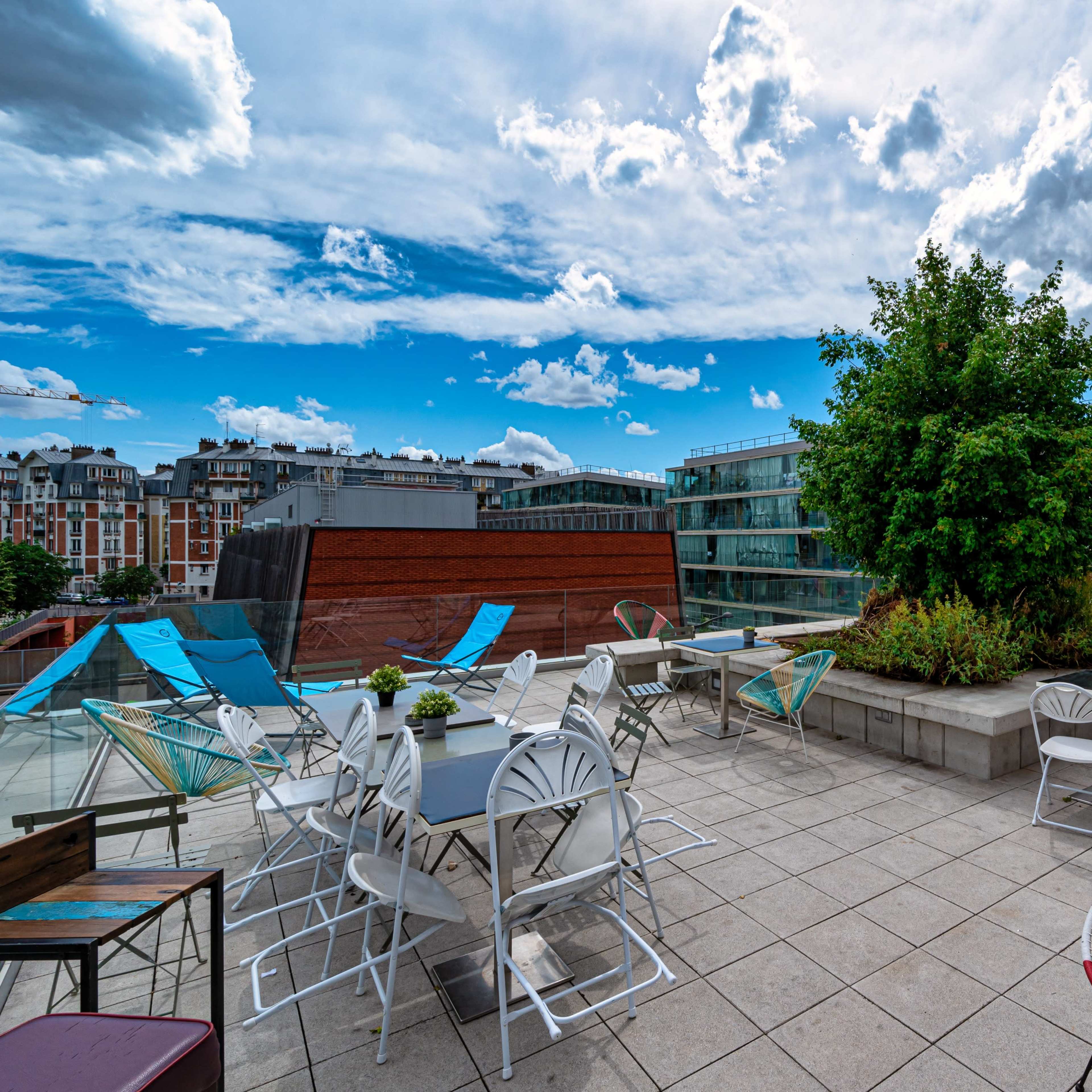 The rooftop terrace features a variety of tables and chairs surrounded by greenery under a cloudy sky.
