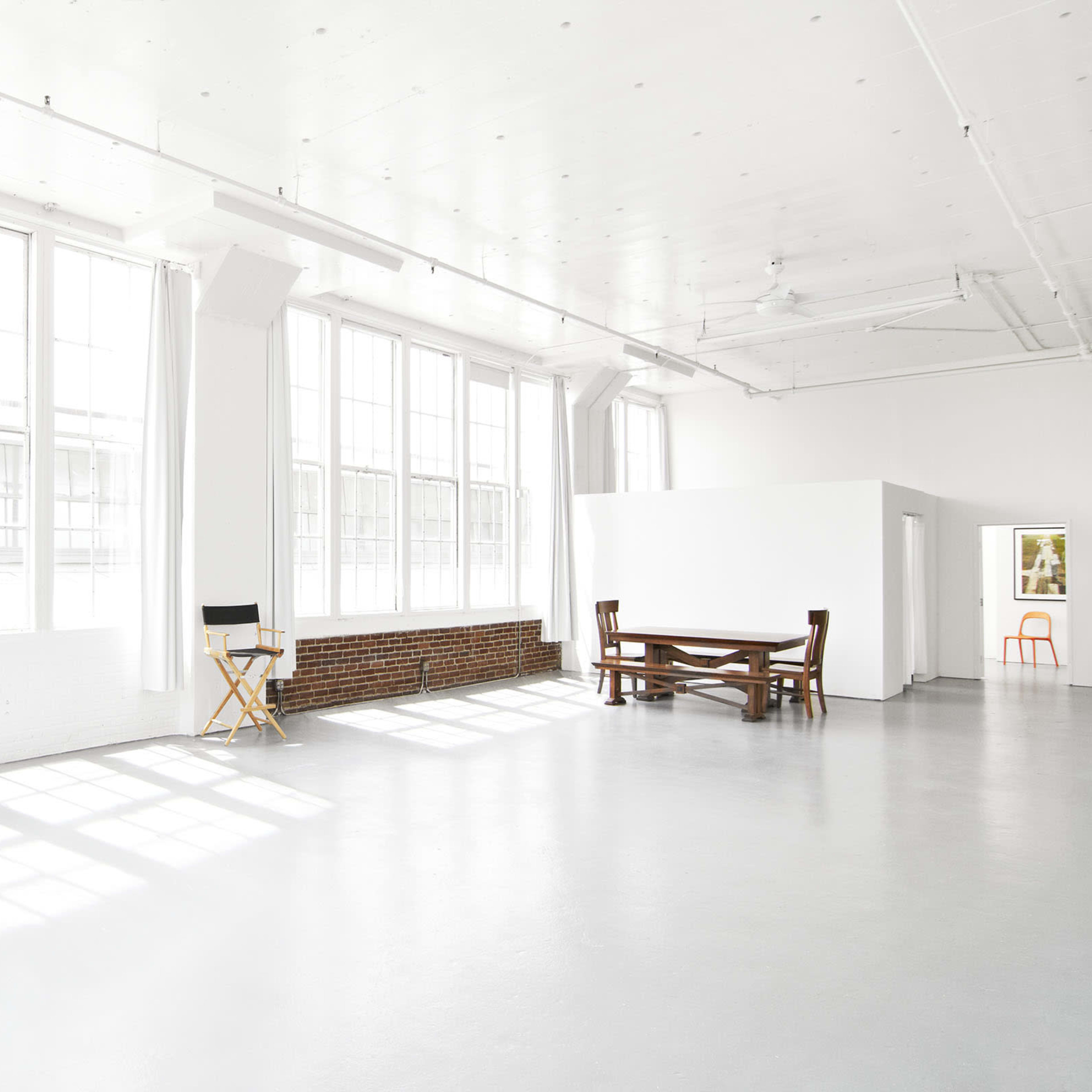 A bright, spacious room with large windows, a wooden table, a director's chair, and a white wall.