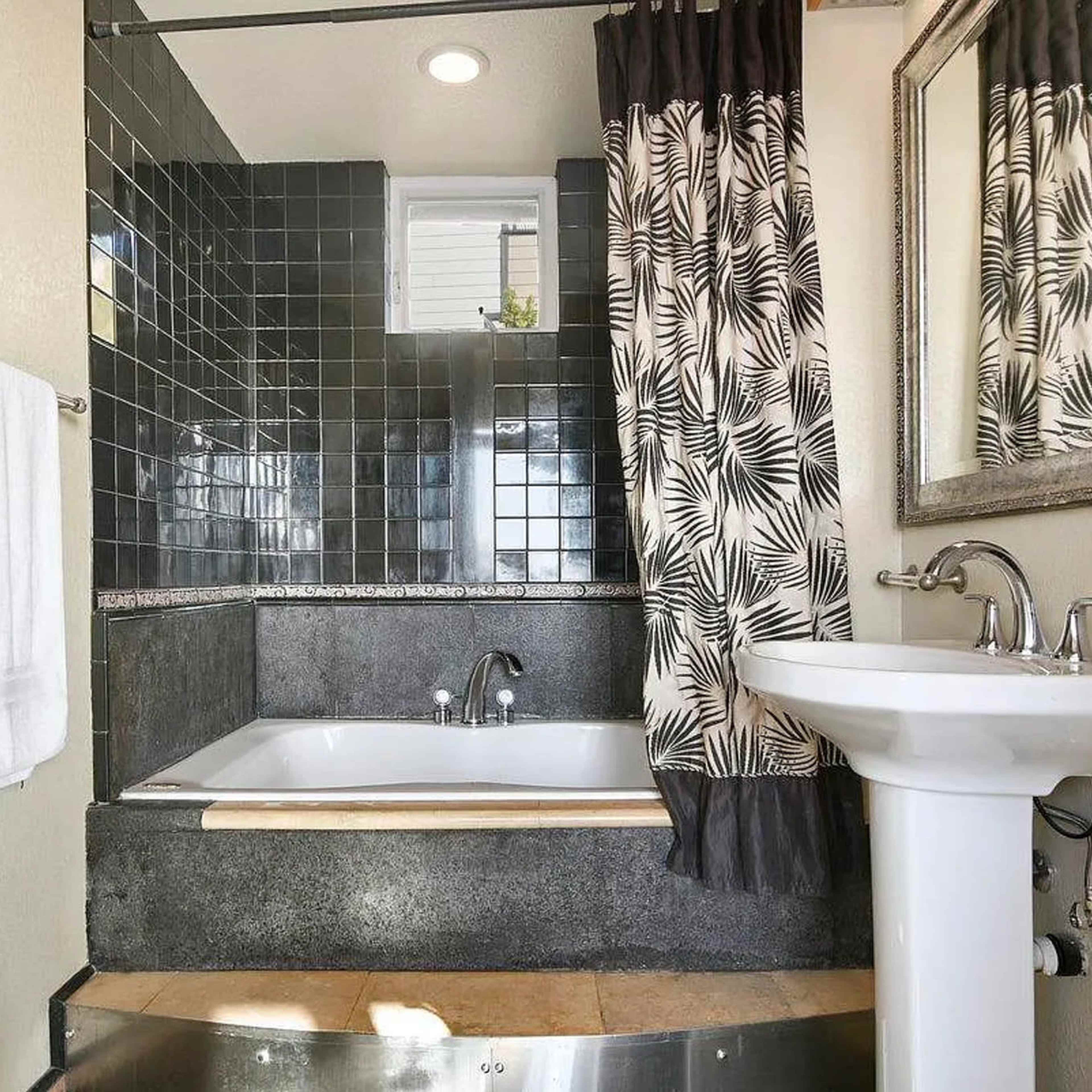 A bathroom featuring a bathtub, a pedestal sink, and black tiled walls with a patterned shower curtain.