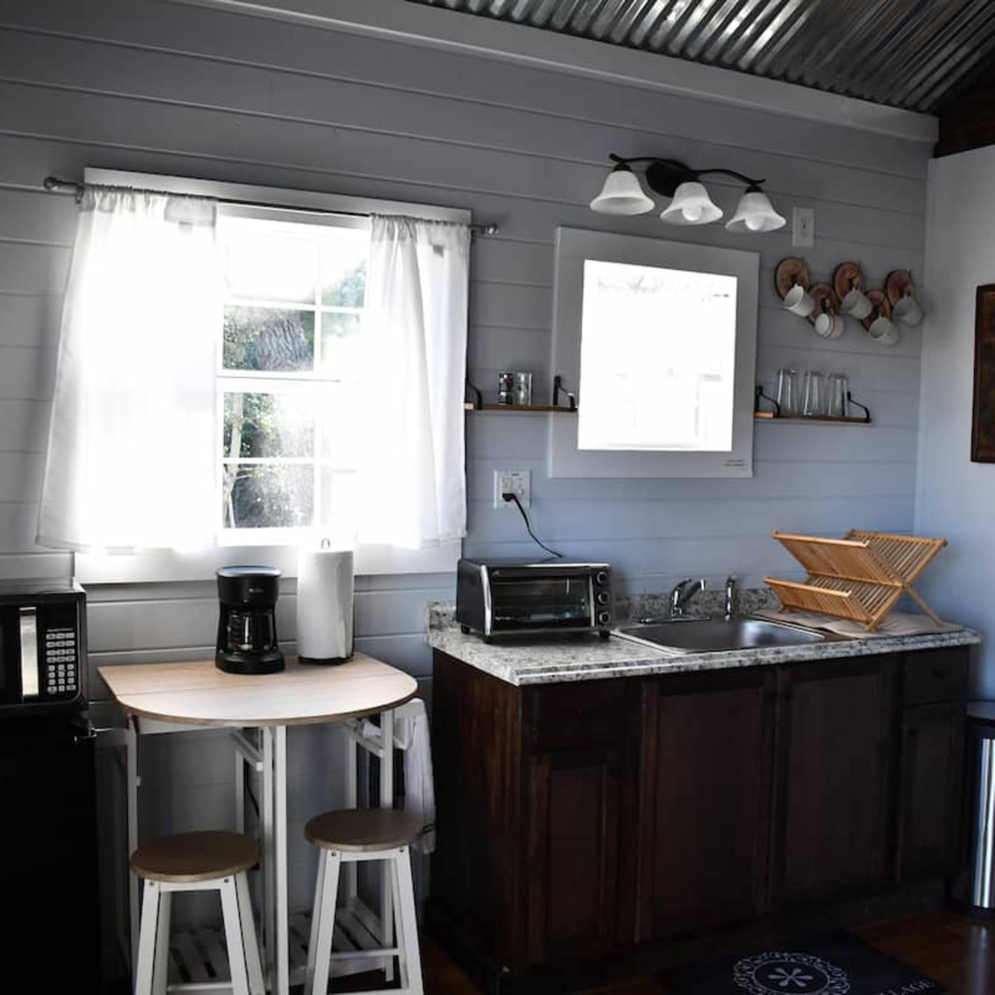 A compact kitchen featuring a countertop with a sink, a microwave, a coffee maker, and a small table with two stools, all against a backdrop of light blue walls and metal accents.