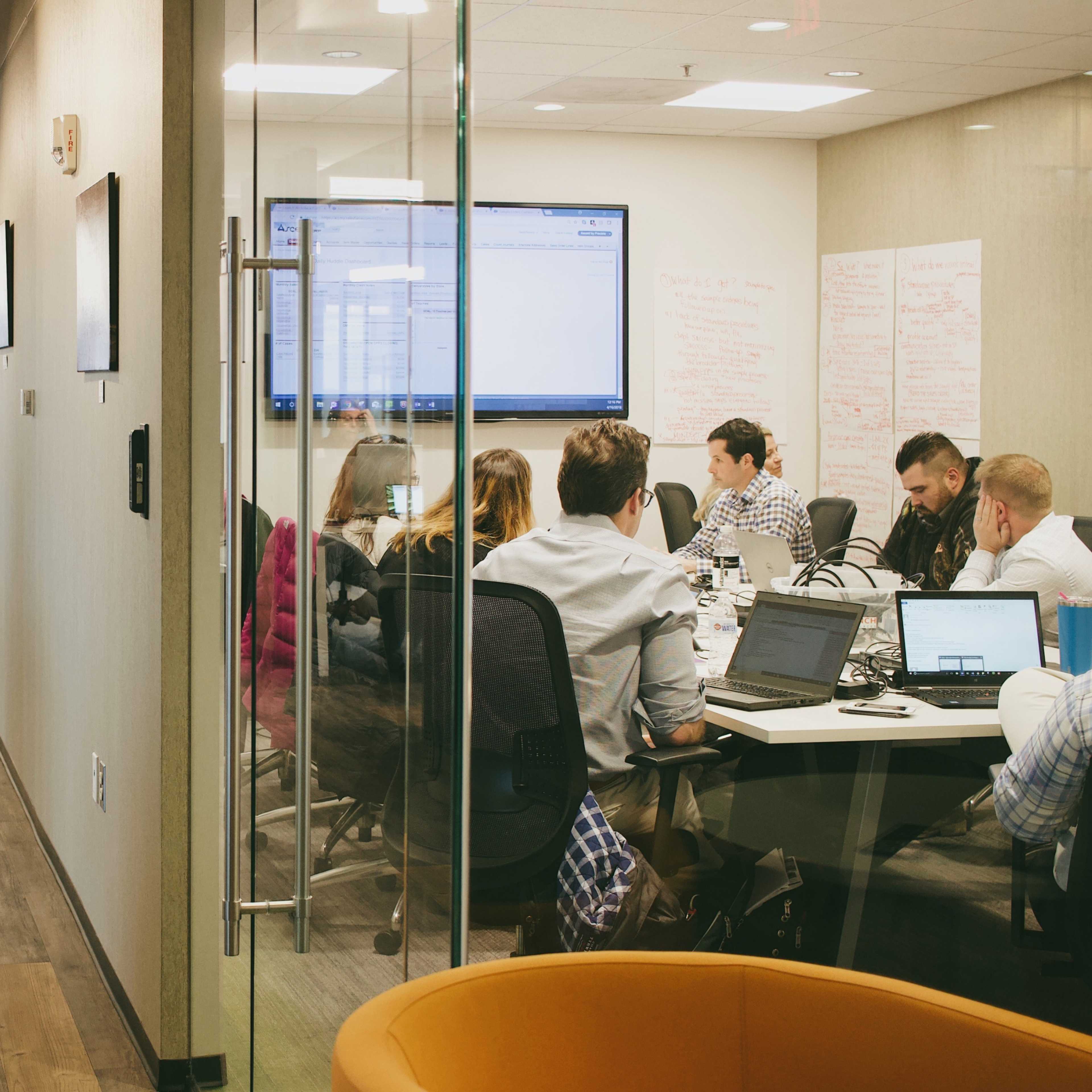 A group of people is seated around a conference table in a modern office, with a large screen displaying information in the background.