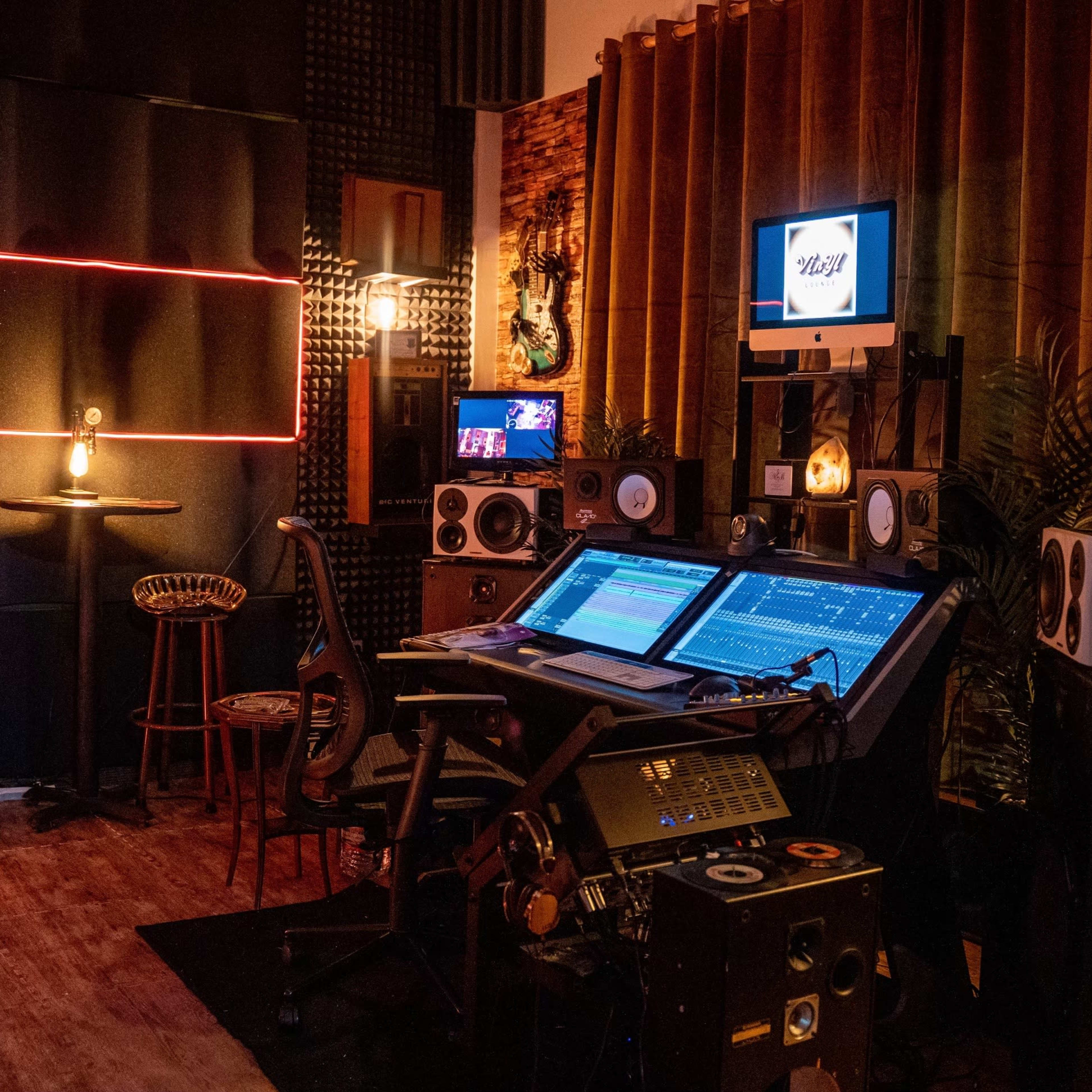 A music studio featuring a large mixing console, speakers, and a cozy seating area with bar stools against a backdrop of acoustic panels.