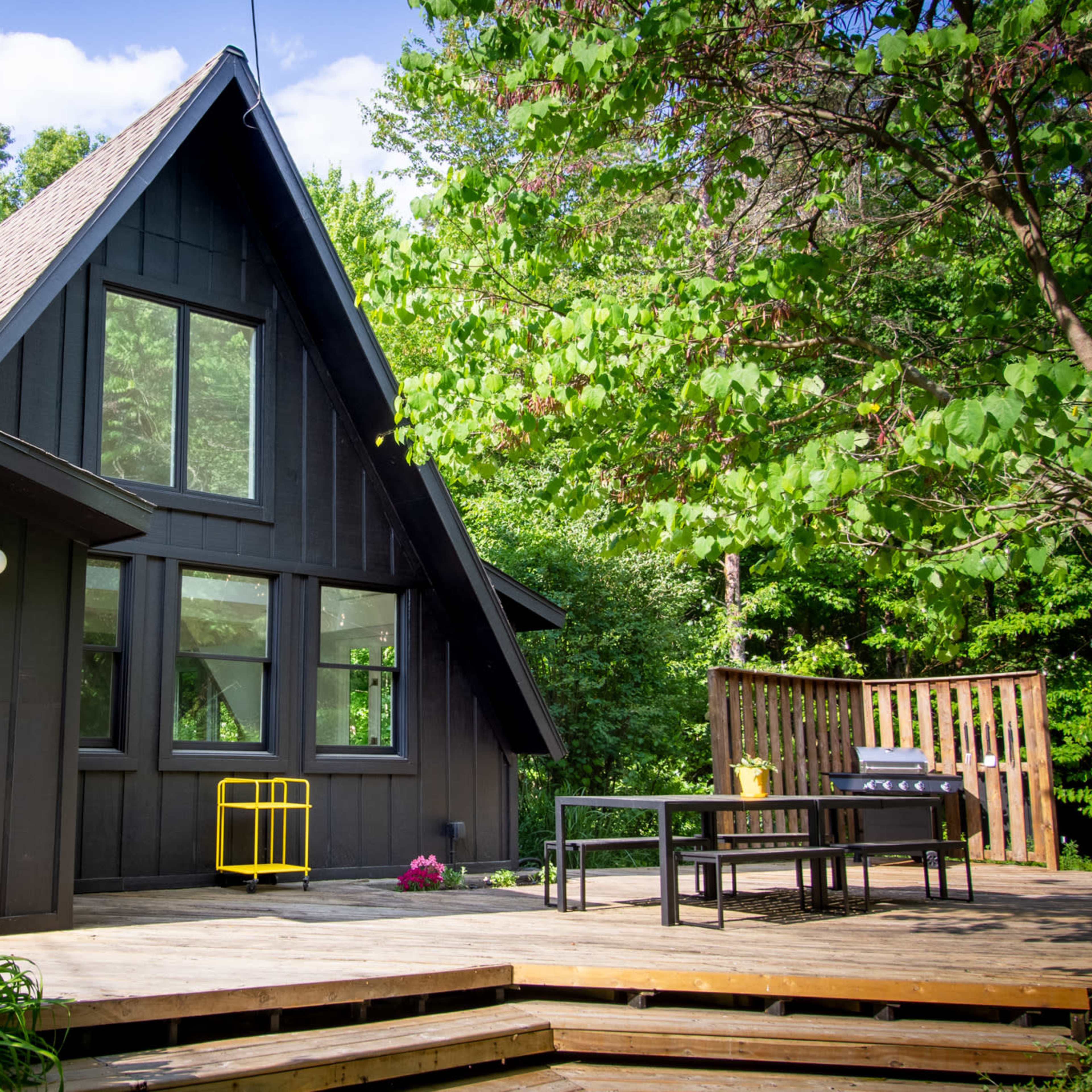 A triangular-roofed black cabin with large windows overlooks a wooden deck surrounded by greenery.