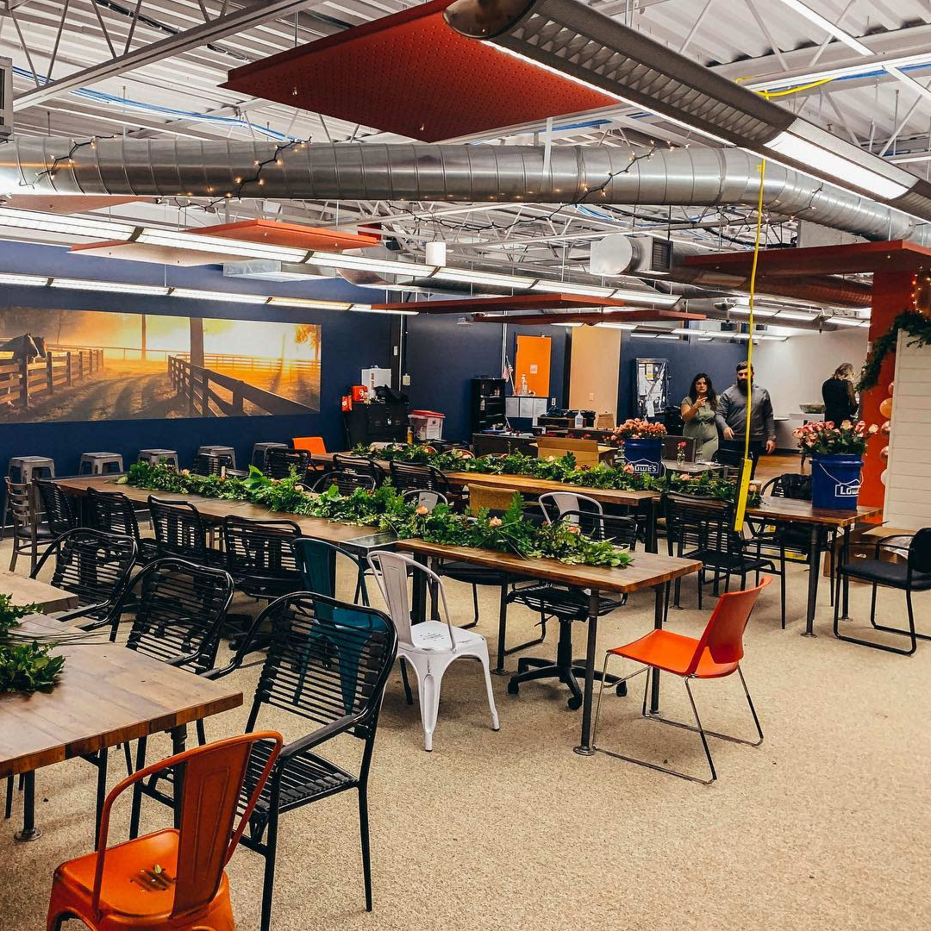 A large, open room is filled with long communal tables and chairs, featuring greenery decor on the tables and a wall mural of a scenic pathway.