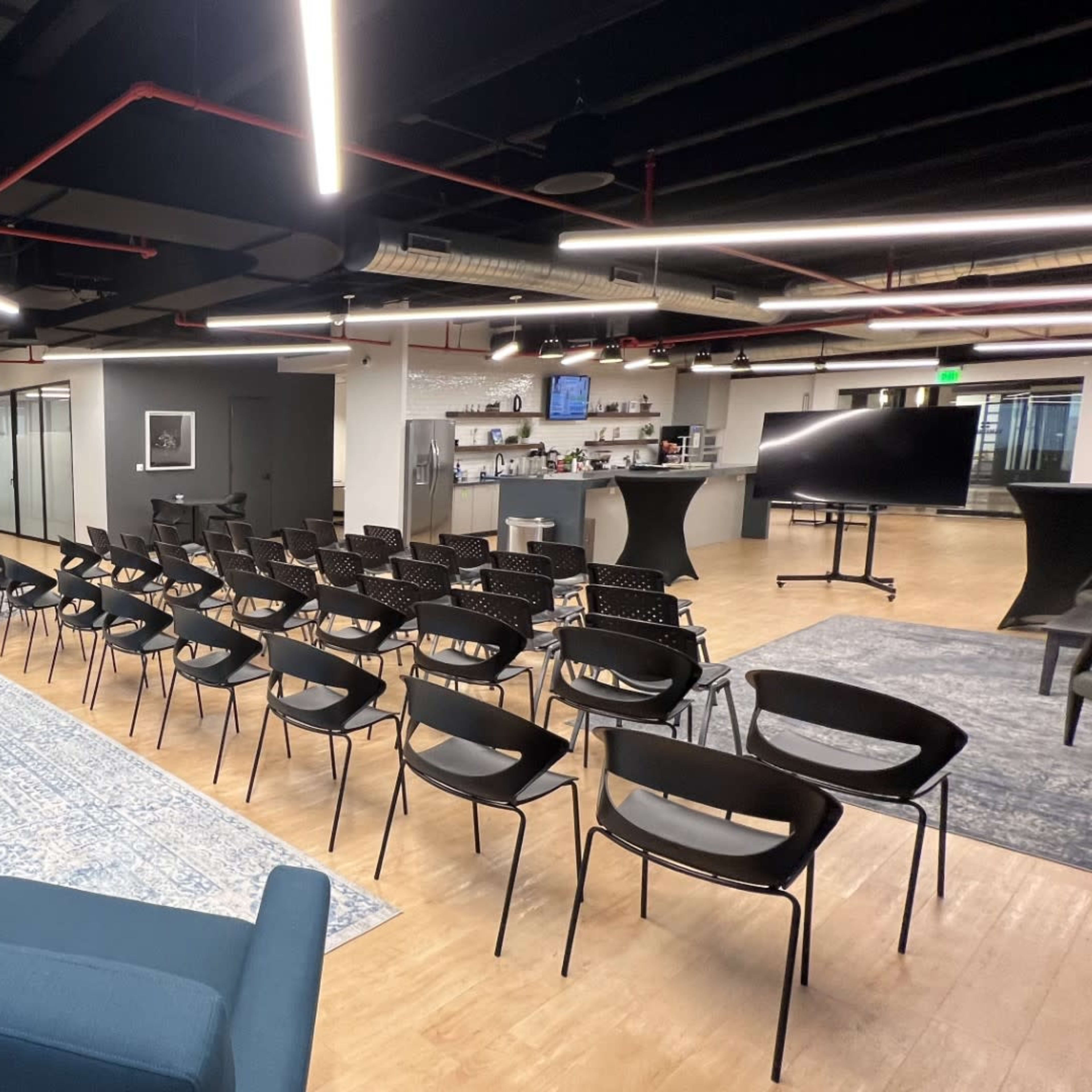 A modern office space with seating areas featuring blue chairs, a conference setup with black chairs facing a presentation area, and a kitchen area in the background.