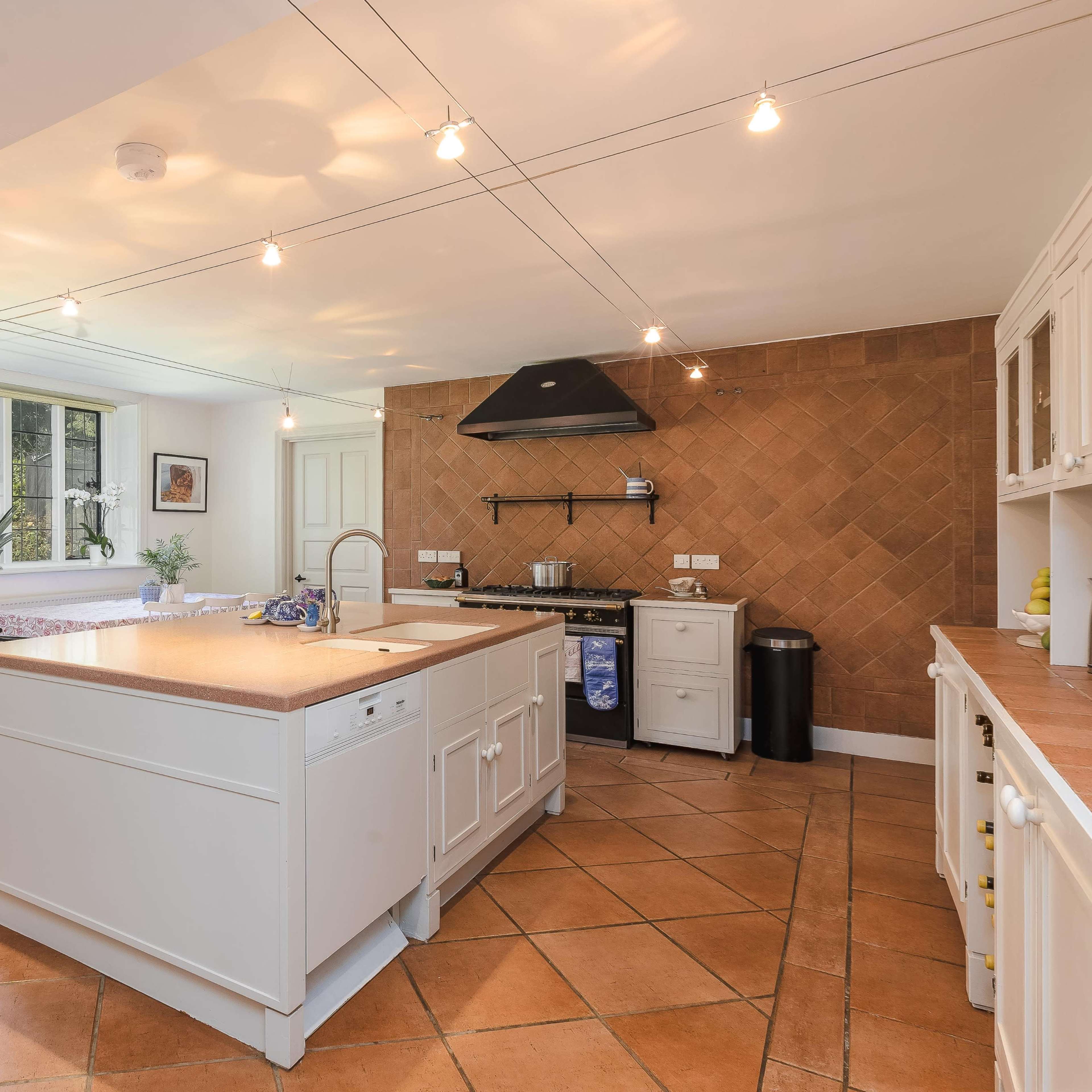 The kitchen features a large island with a sink, surrounded by light-colored cabinetry and a warm tiled floor, with natural light streaming in through large windows.