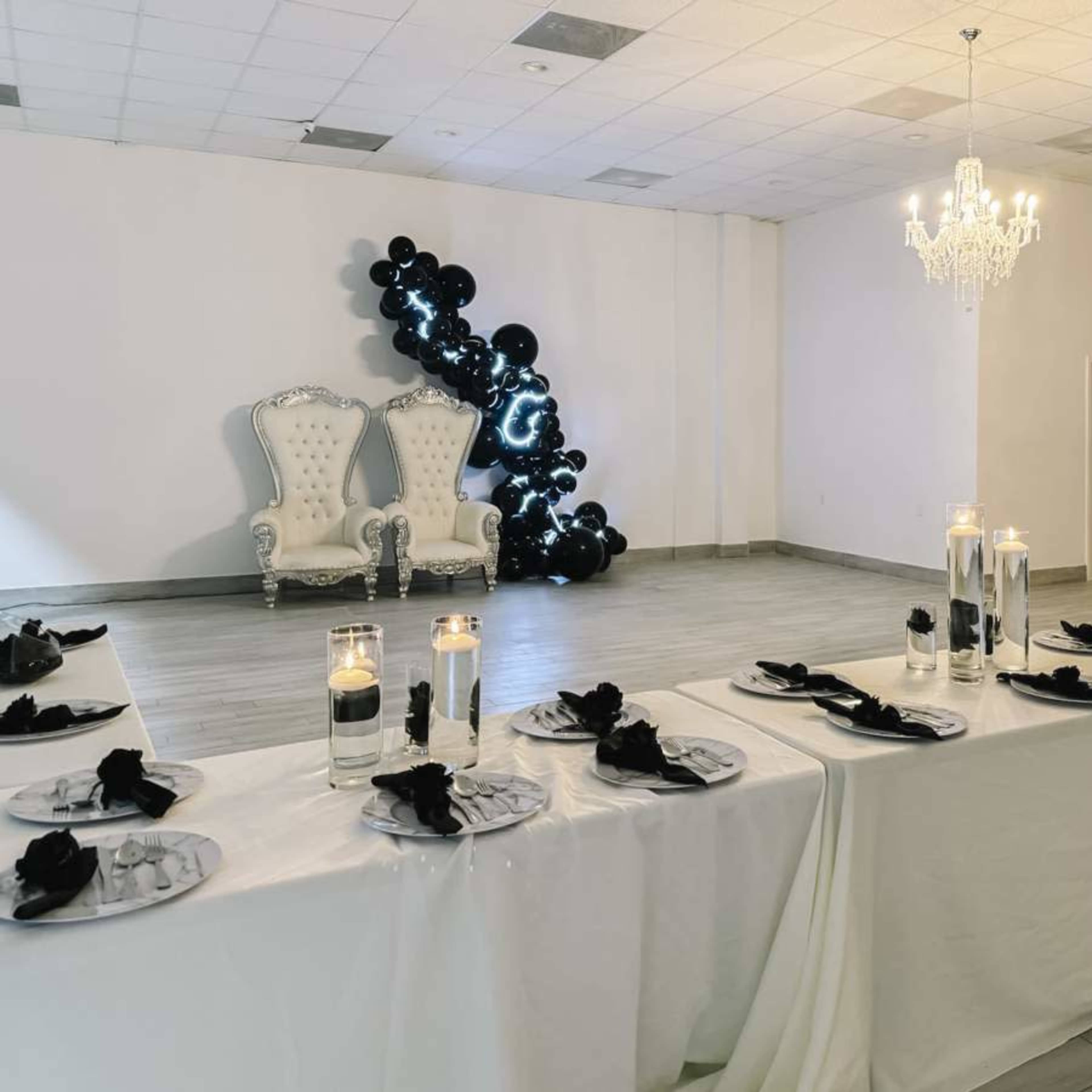 A beautifully arranged banquet hall with a long table set for a formal event, featuring white tablecloths, black napkins, and a decorative backdrop of balloons, alongside an ornate white throne chair and a chandelier.