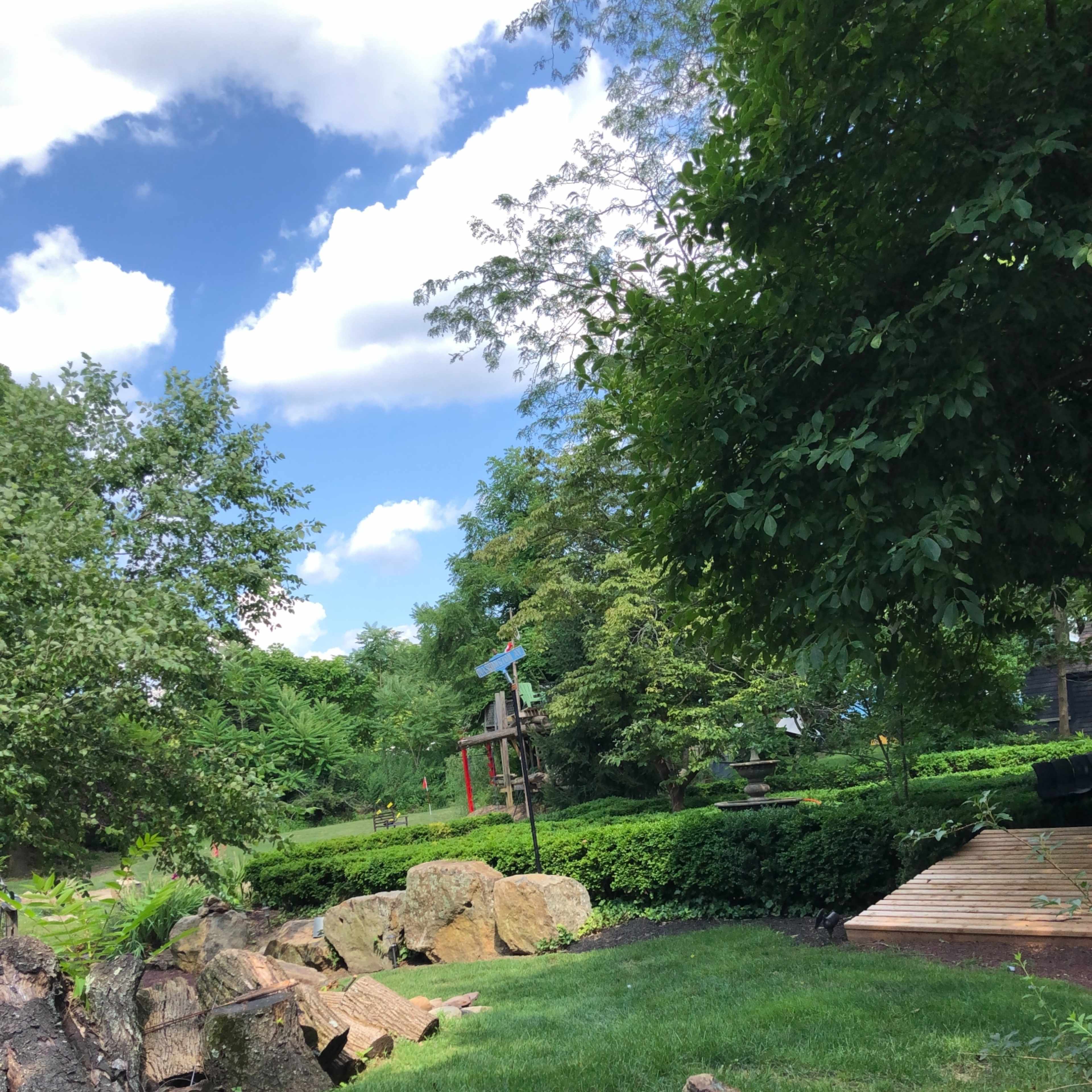 A green backyard with rocks, a wooden deck, and a playground set in the distance under a partly cloudy sky.