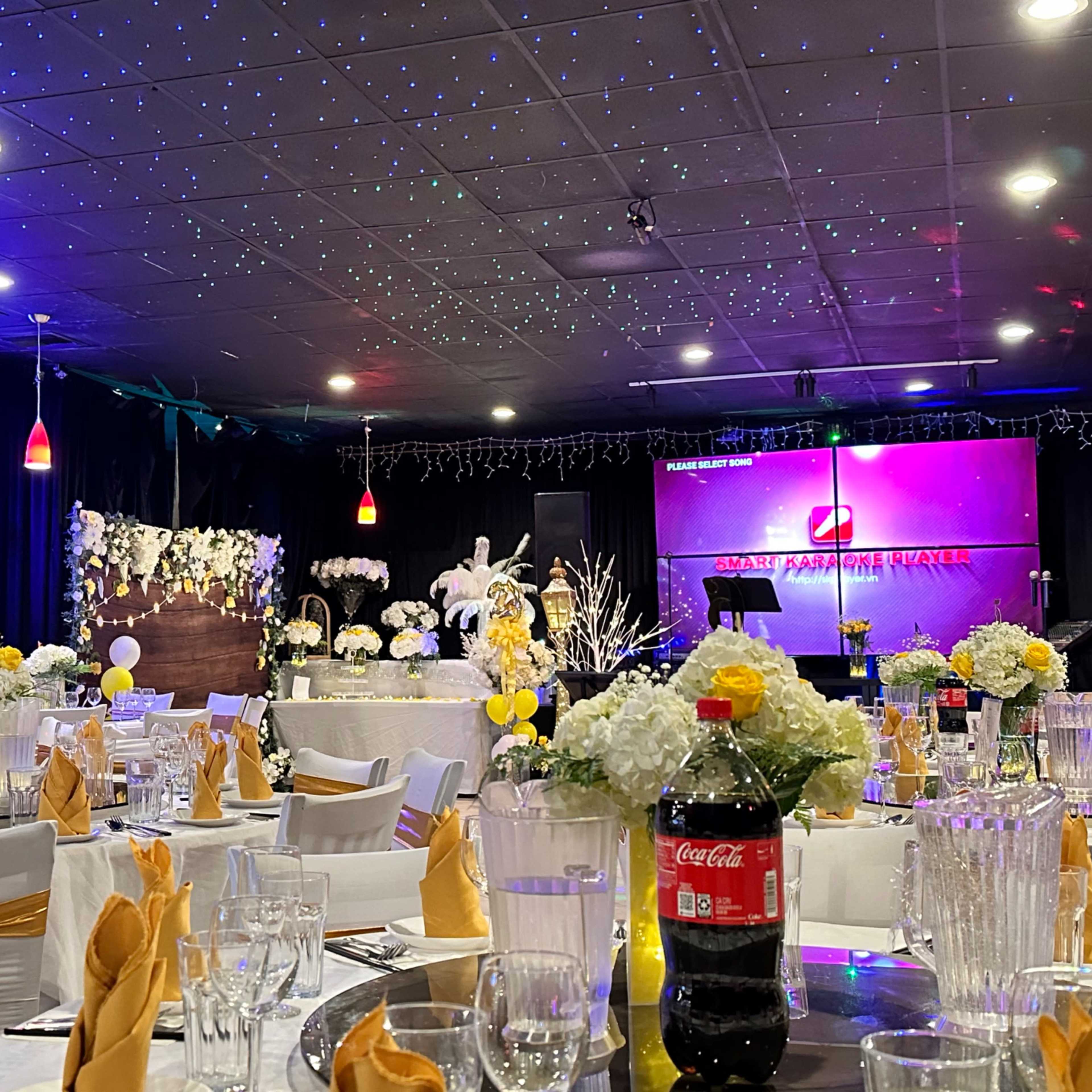 A decorated banquet hall with tables set for a celebration, featuring white and yellow floral arrangements and a stage with colorful lighting.