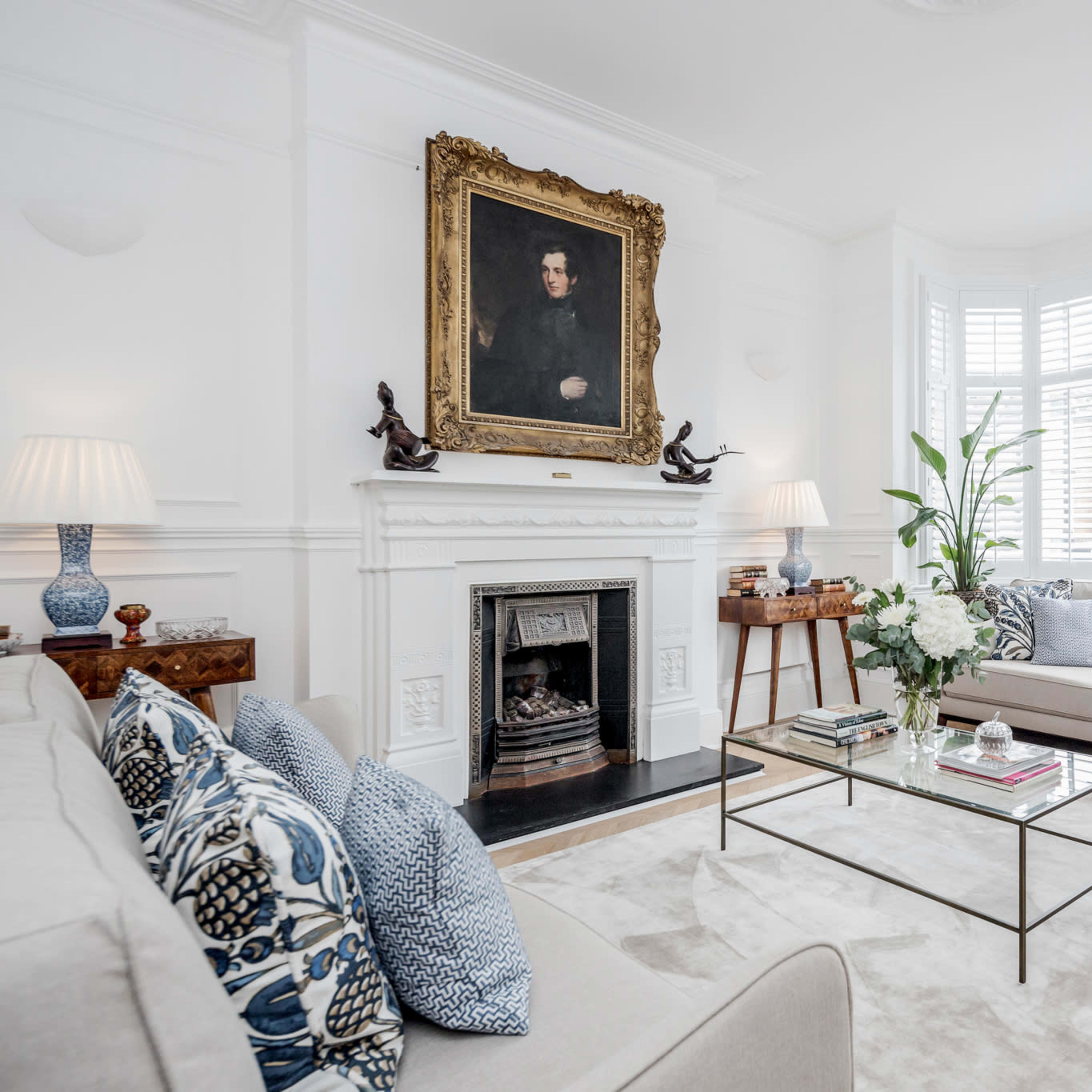 A bright living room features a fireplace, a large portrait above the mantel, a plush sofa with decorative pillows, and a glass coffee table surrounded by elegant decor.
