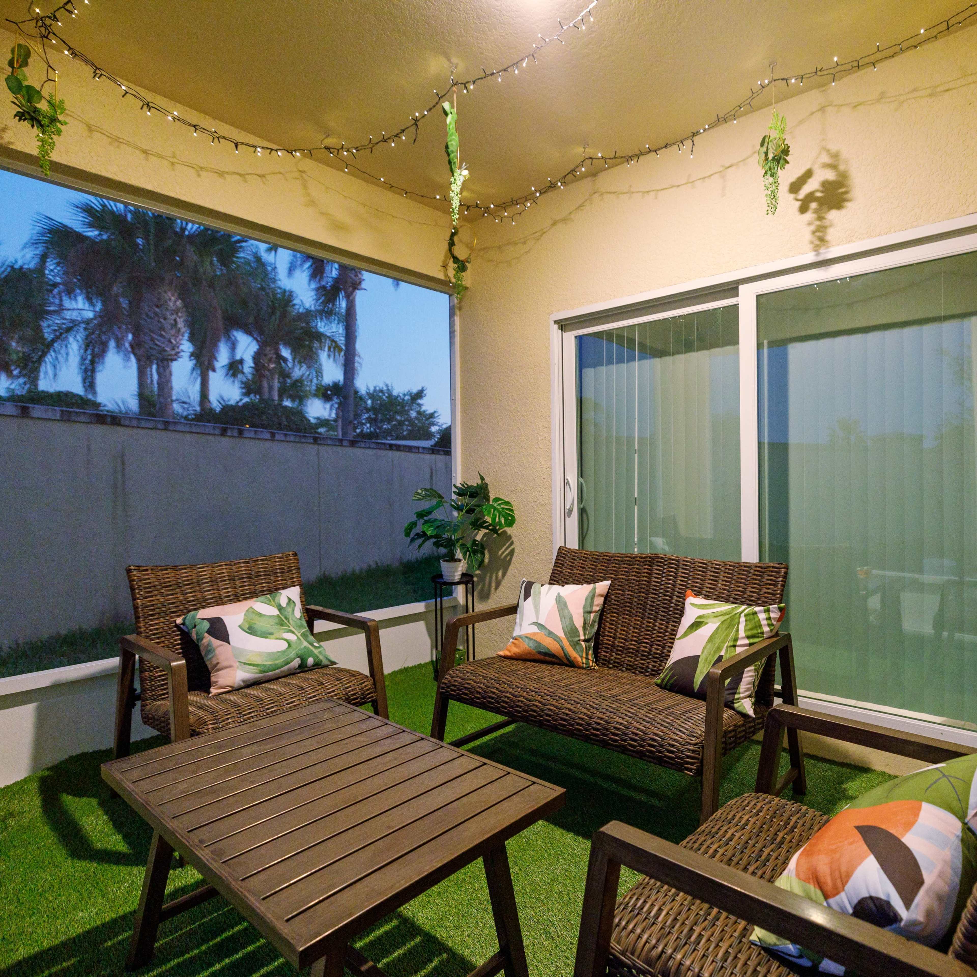 A cozy outdoor patio features a table surrounded by wicker seating, accent pillows, and potted plants, with sliding glass doors leading to a view of palm trees.
