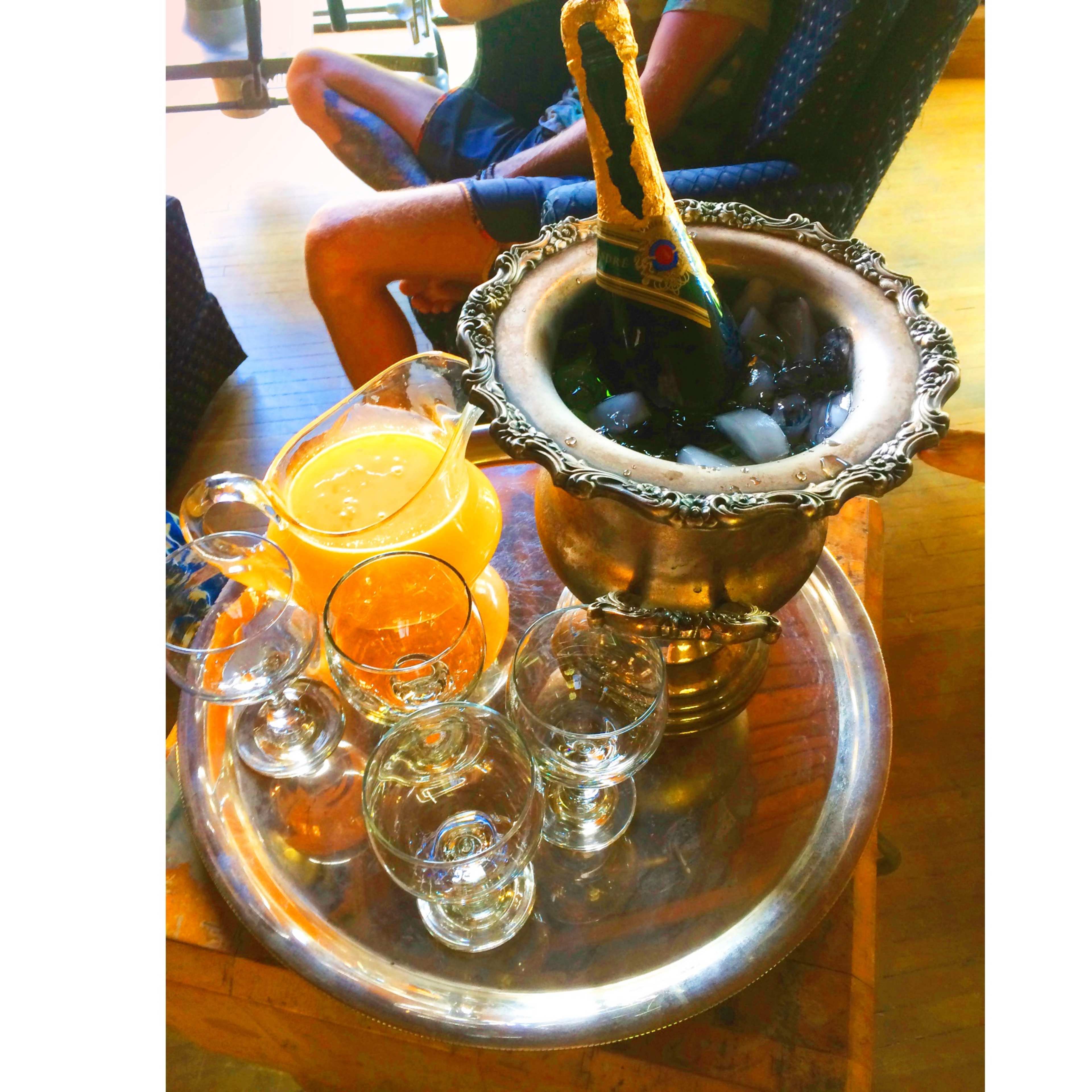 A silver ice bucket filled with ice holds a bottle of champagne, accompanied by various glassware and a pitcher of orange juice on a serving tray.