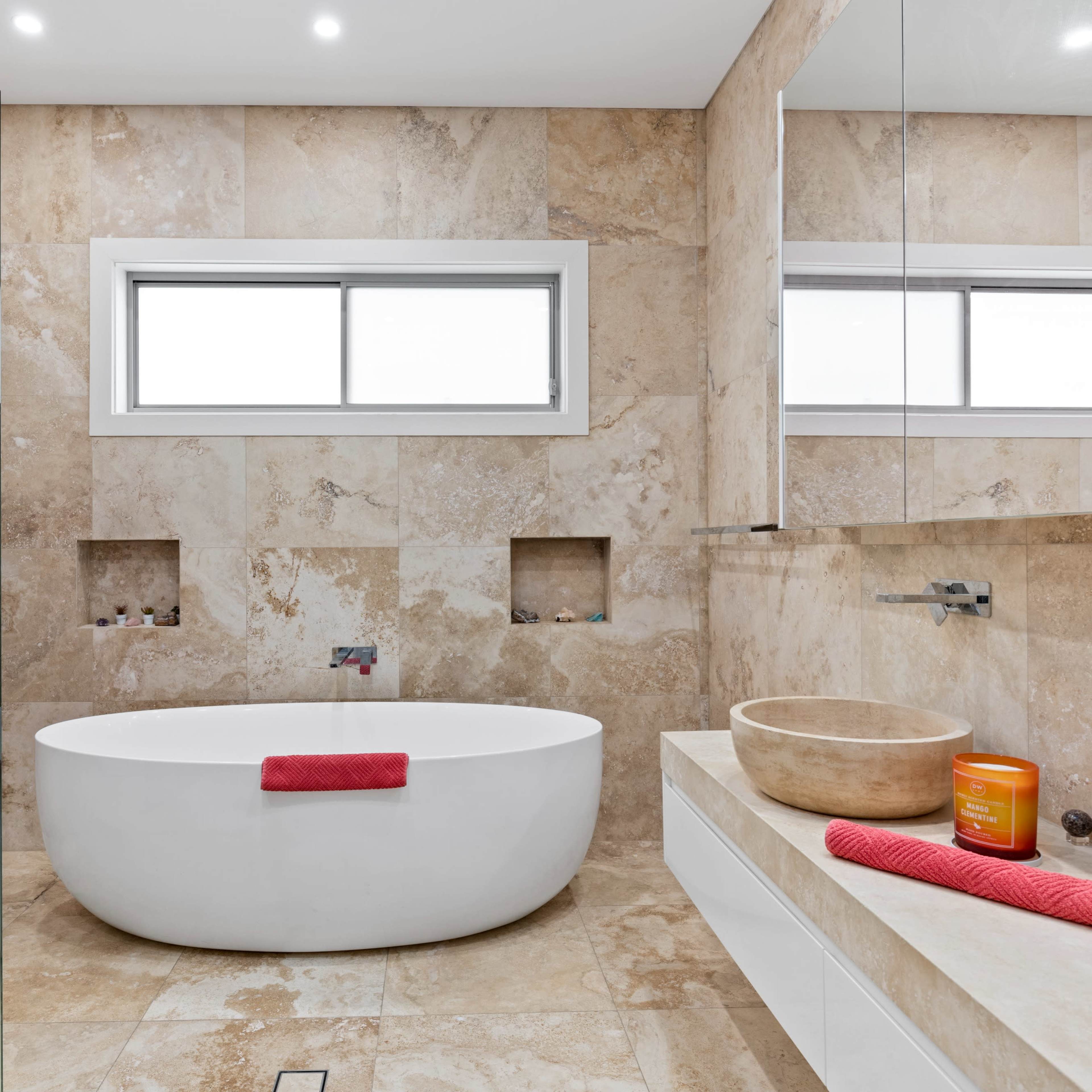 The bathroom features a freestanding white bathtub, a large mirror, and beige stone walls with two built-in niches.