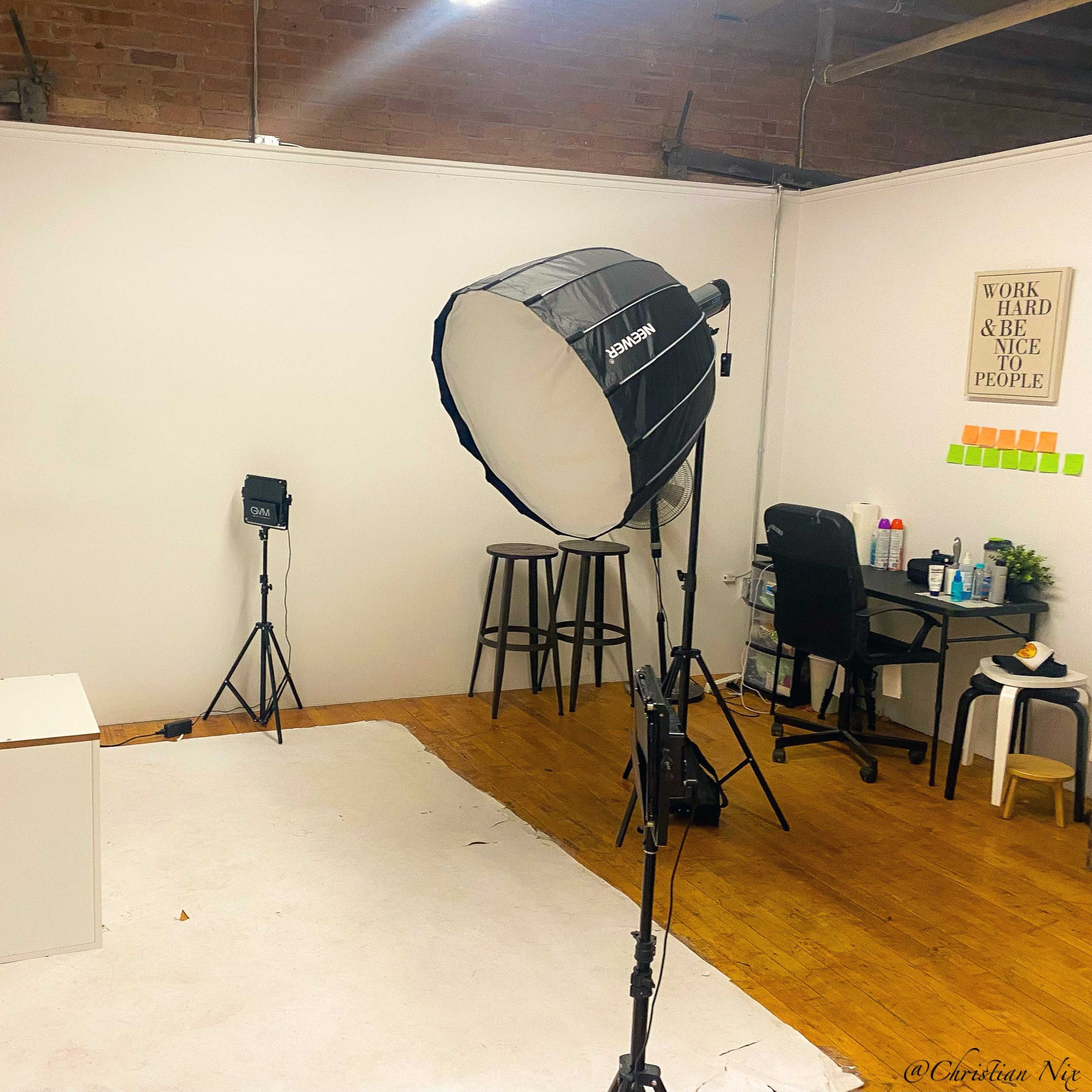 A photography studio with a light umbrella, two stand lights, a stool, and a chair against a plain wall.