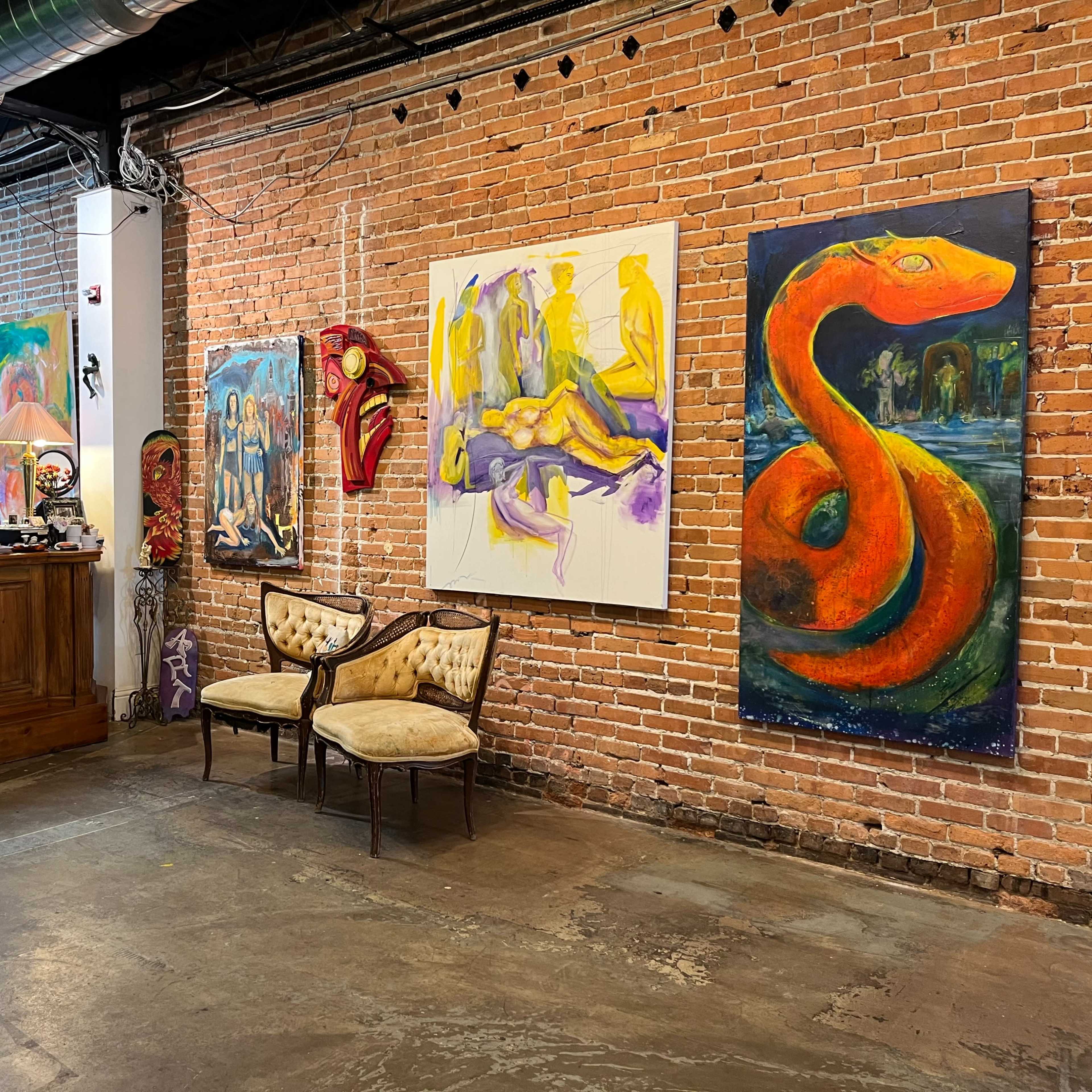 An art gallery with exposed brick walls featuring colorful paintings, a wooden reception desk, and two upholstered chairs.