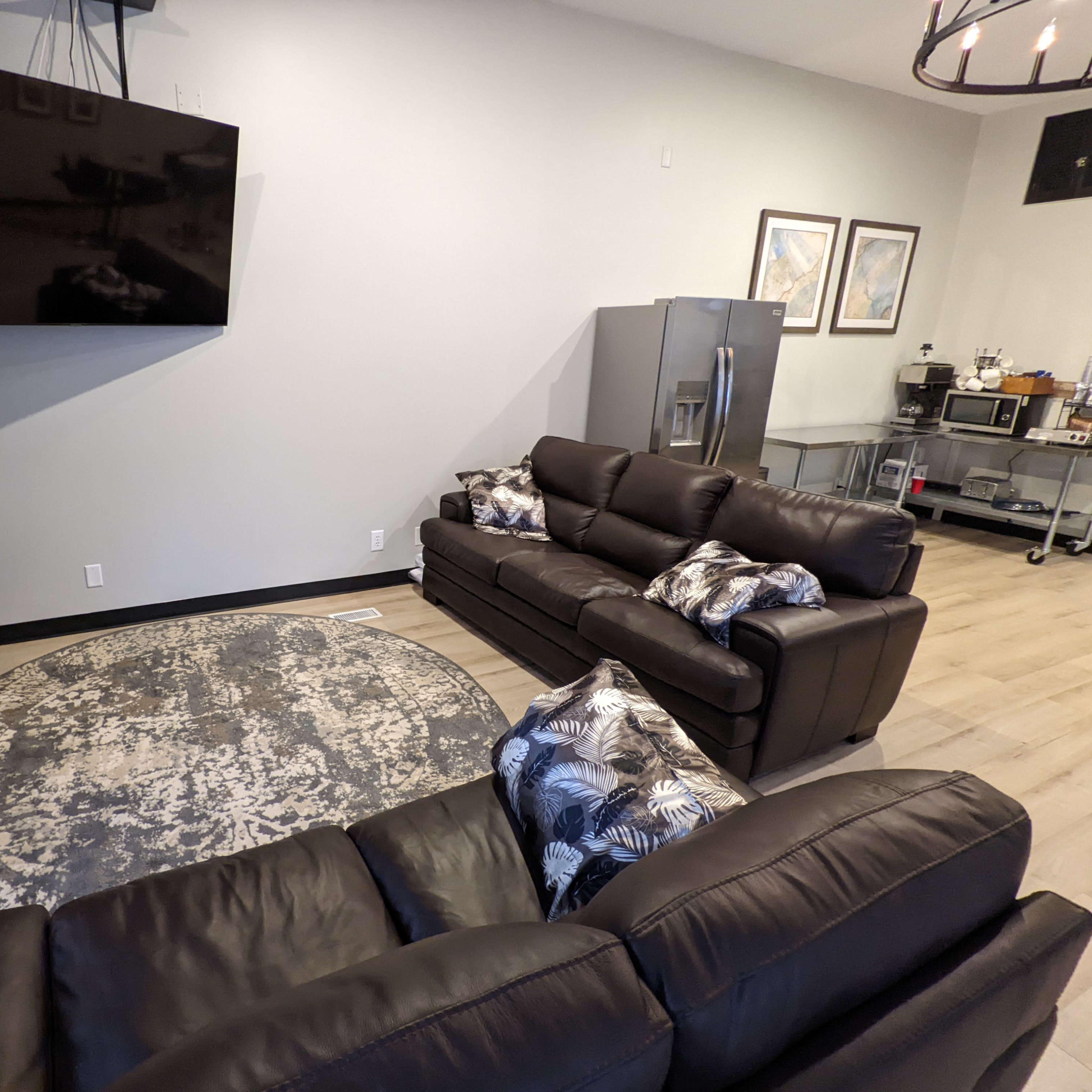 A modern living room with two brown sofas, a round area rug, a wall-mounted television, and a stainless steel refrigerator in the background.