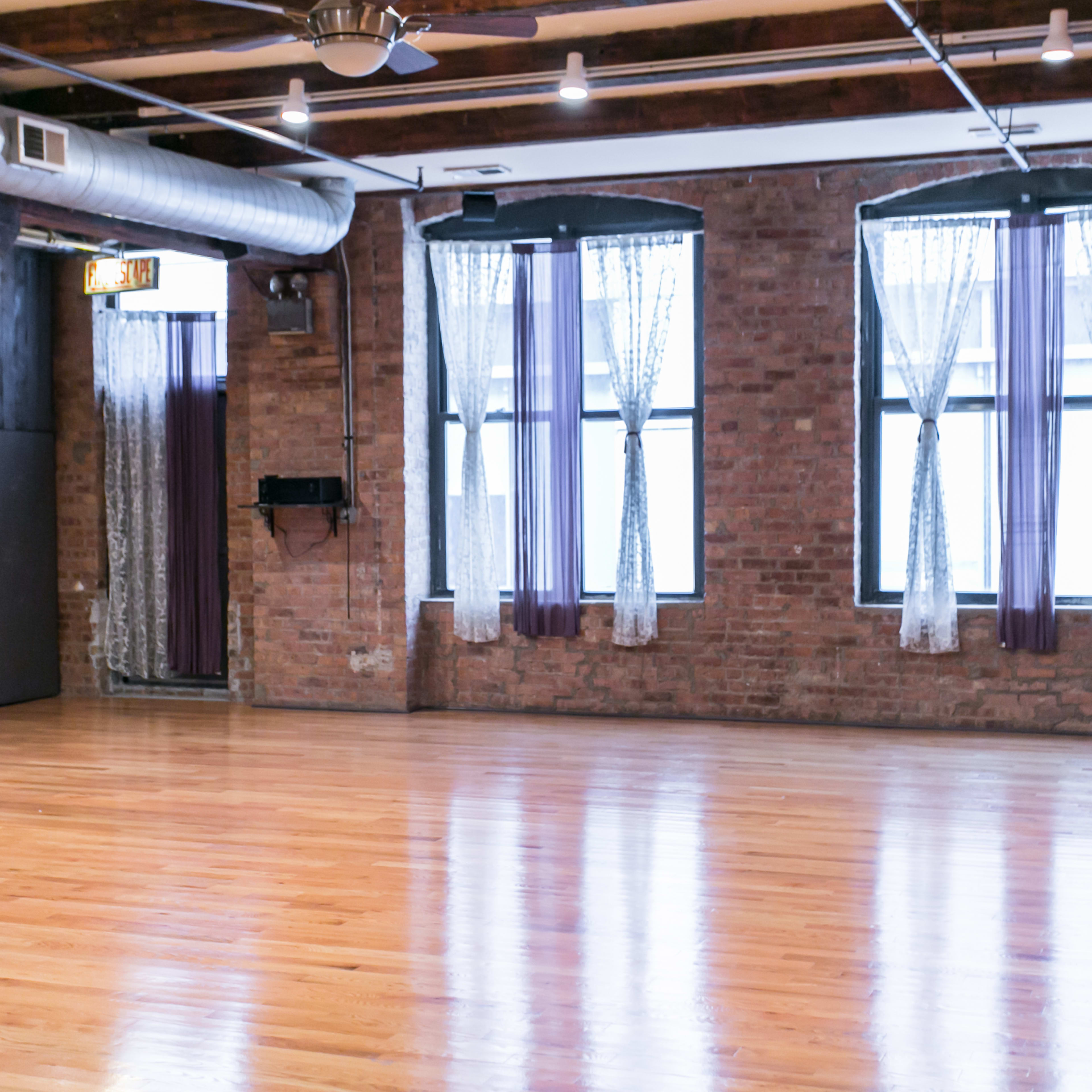 A spacious room with hardwood flooring, exposed brick walls, large windows with sheer curtains, and wooden beams in the ceiling.
