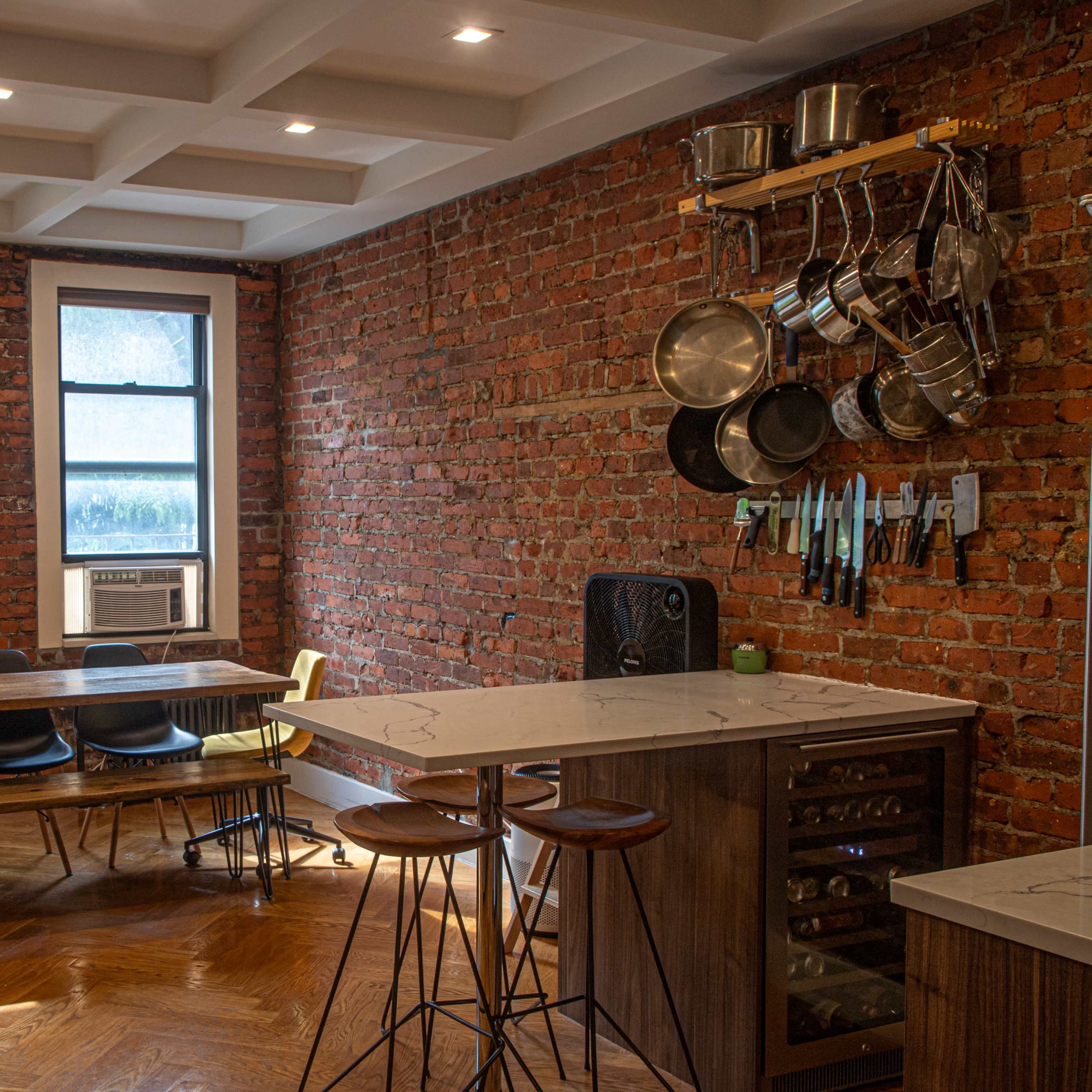 A modern kitchen features exposed brick walls, a large wooden table, and stainless steel cookware hanging above the countertop.