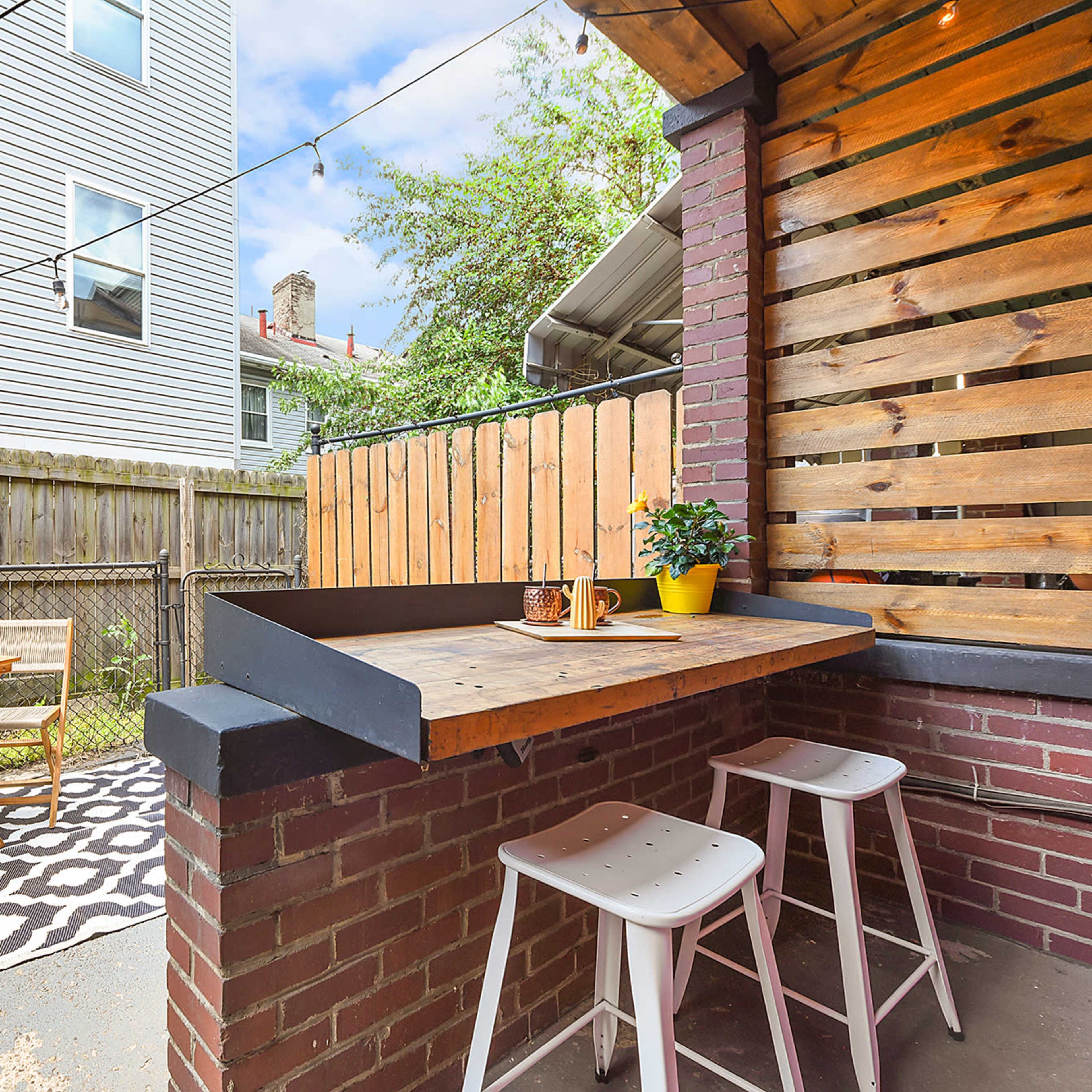 A small outdoor patio area featuring a wooden bar with stools, a table and chairs, and string lights overhead, surrounded by a fenced yard.