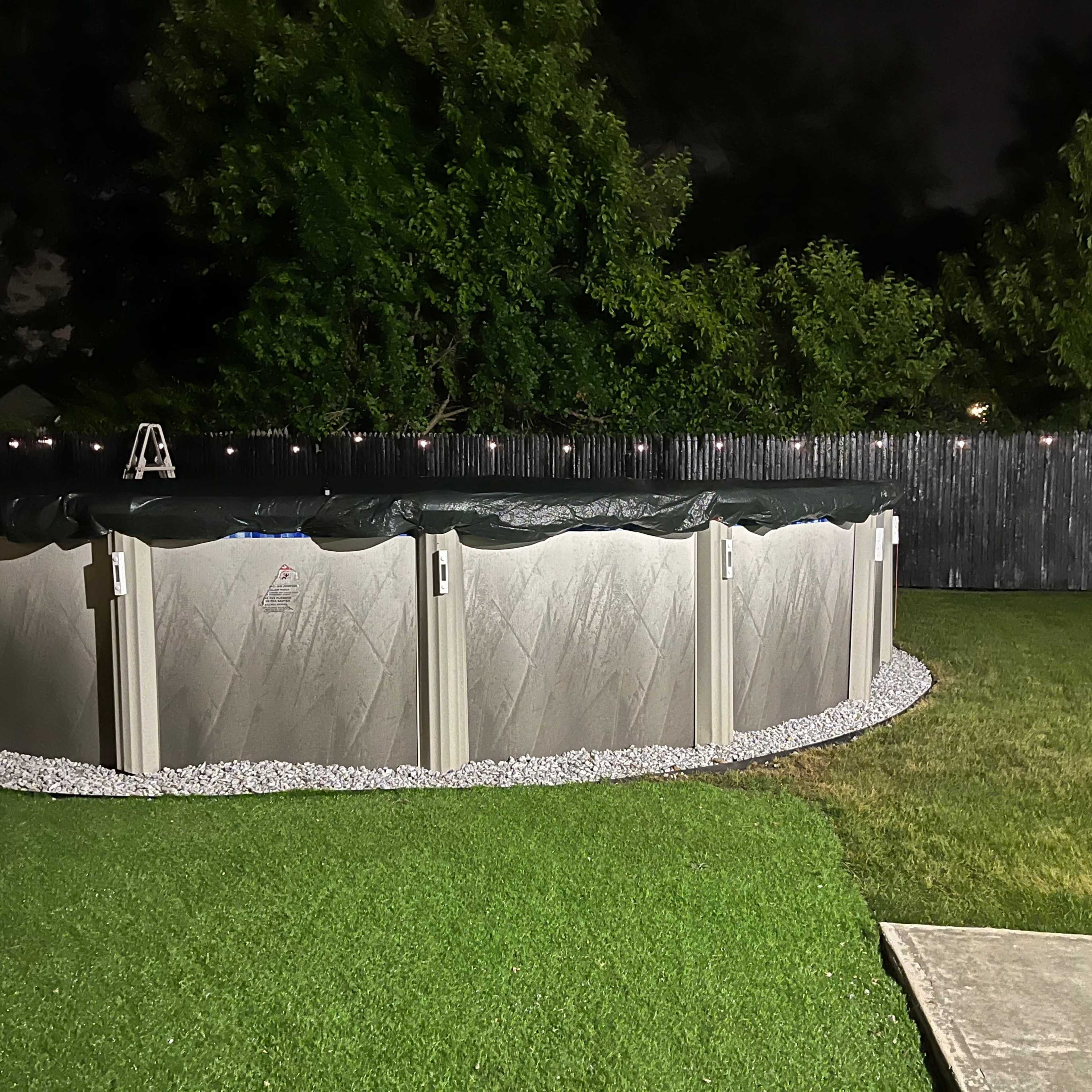 A round above-ground pool is surrounded by gravel and sits in a backyard at night, with a wooden fence and trees in the background.