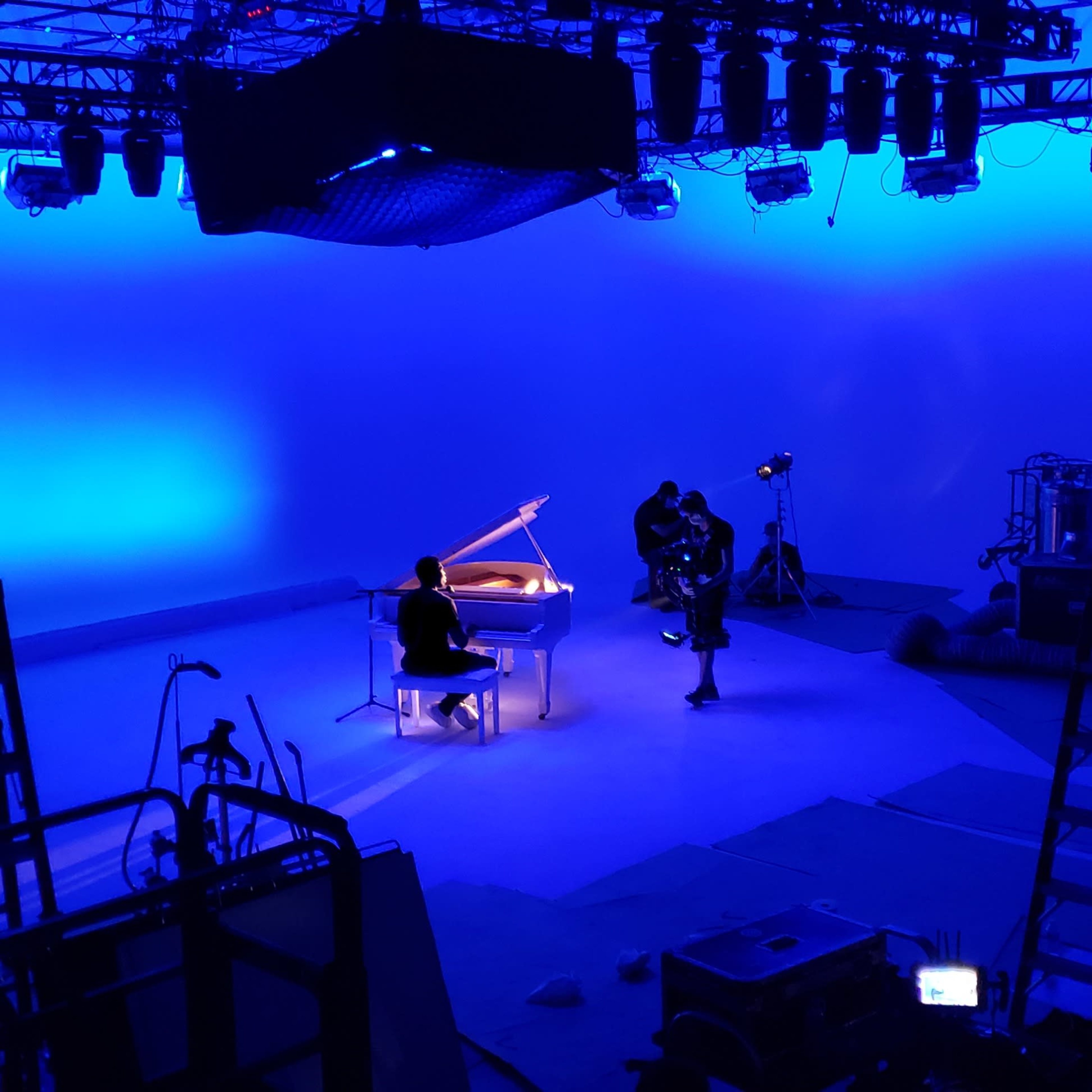 A music studio is set up with a grand piano in the center, illuminated by blue lighting, surrounded by cameras and various equipment.