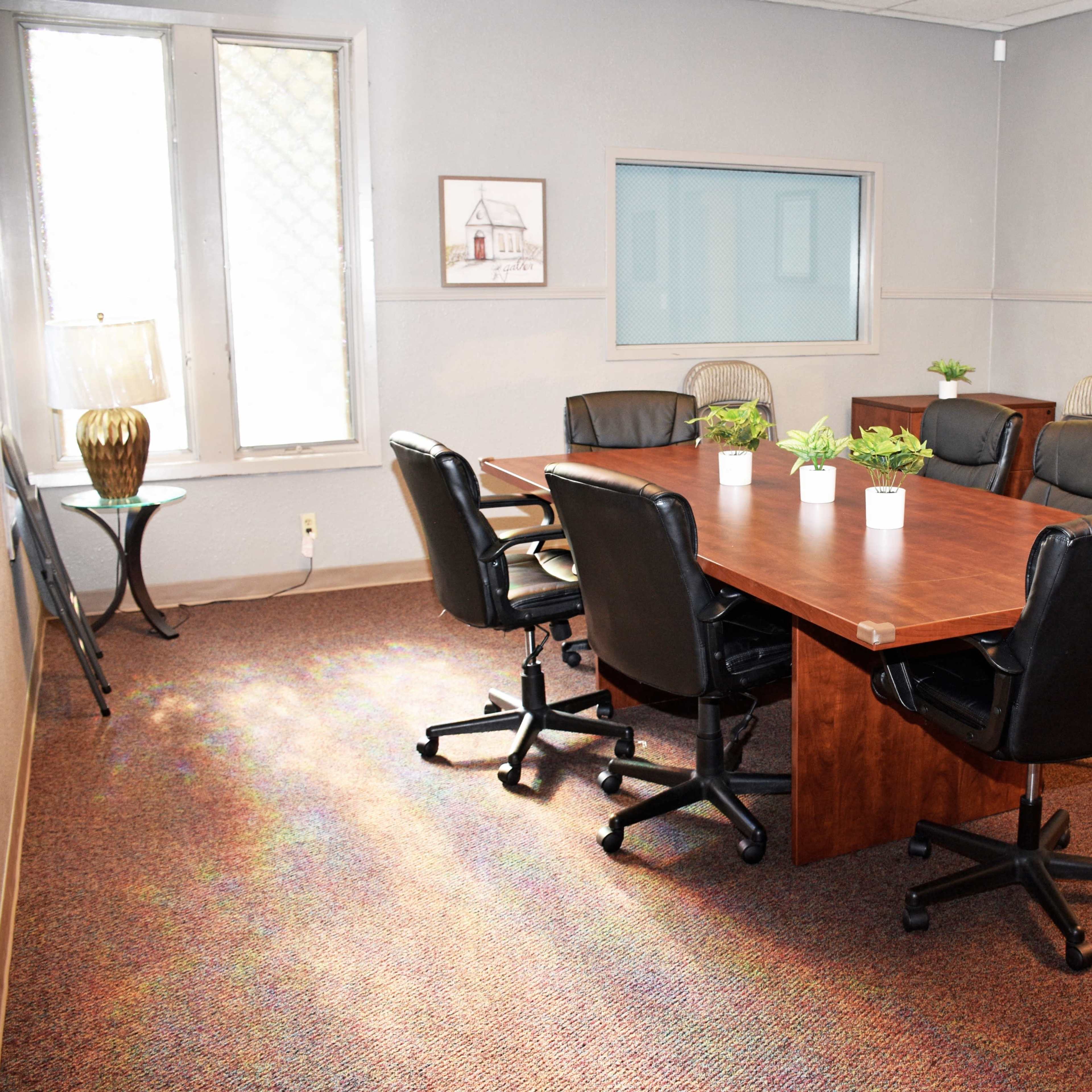 A conference room features a large wooden table surrounded by black rolling chairs, with additional folding chairs positioned against the wall.