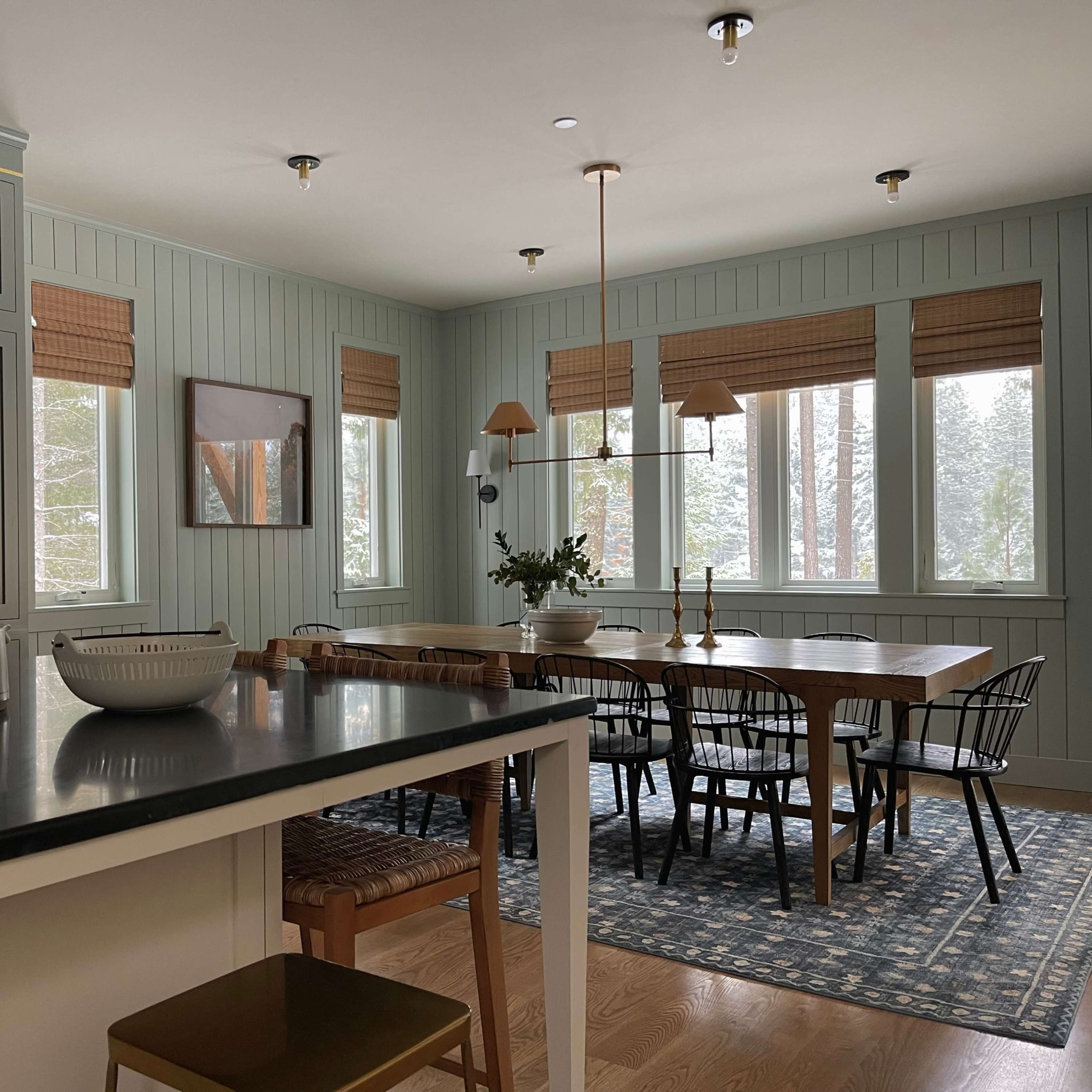 A modern kitchen and dining area featuring a dark countertop, light blue walls, large windows with shades, and a wooden dining table surrounded by black chairs.