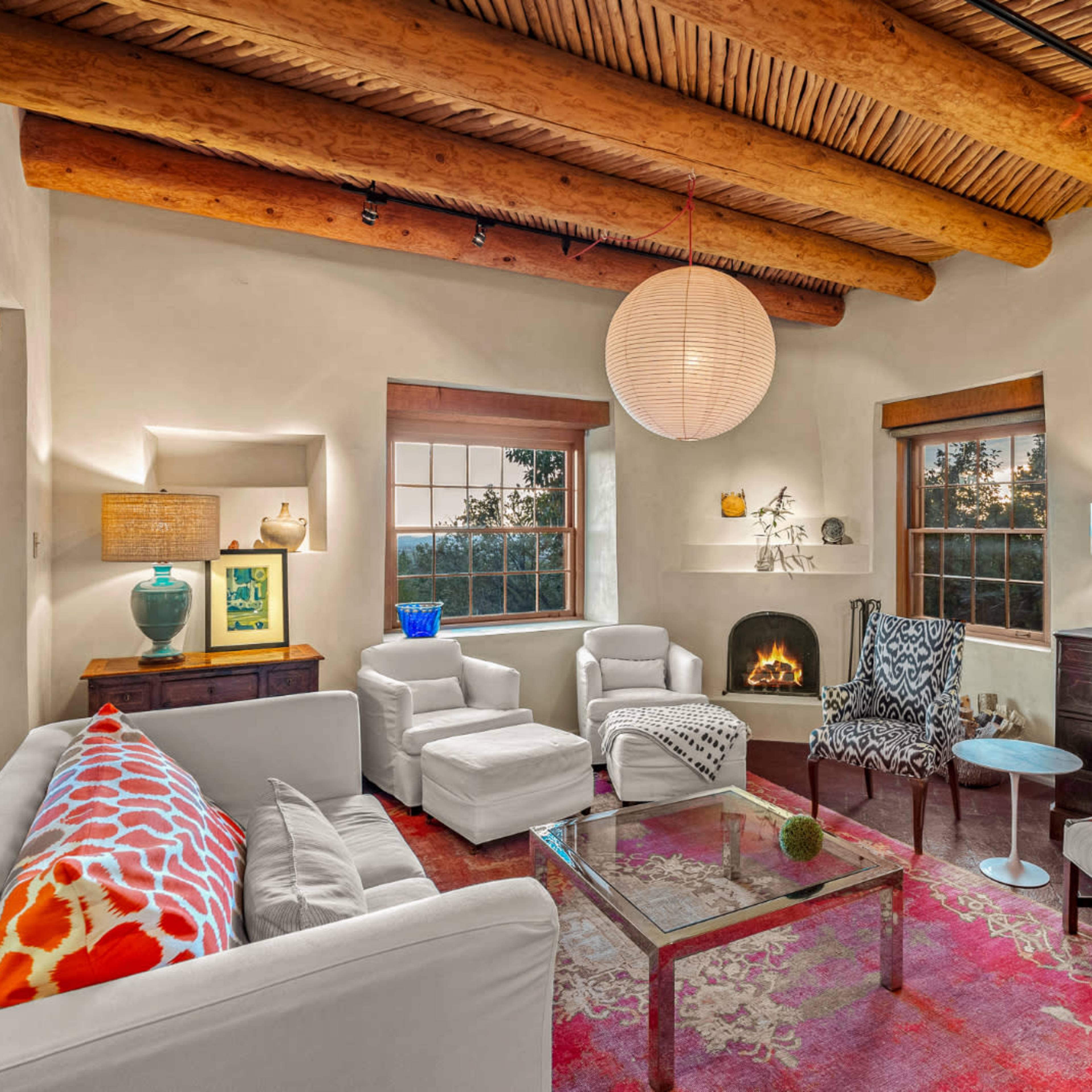 A cozy living room features a mix of white furniture and colorful accents, with wooden beams on the ceiling and large windows providing natural light.