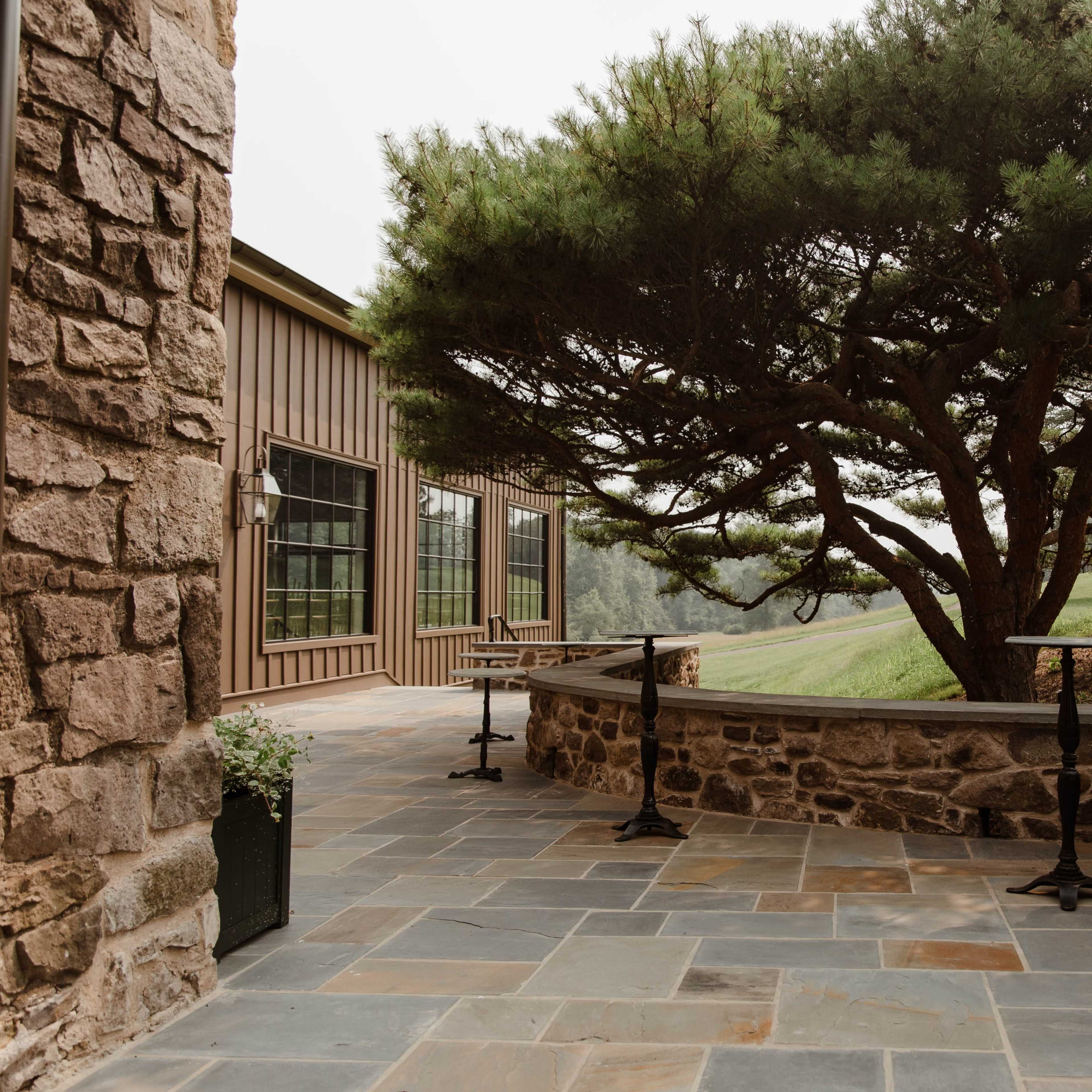 A stone patio is surrounded by a large tree and features two black bistro tables.