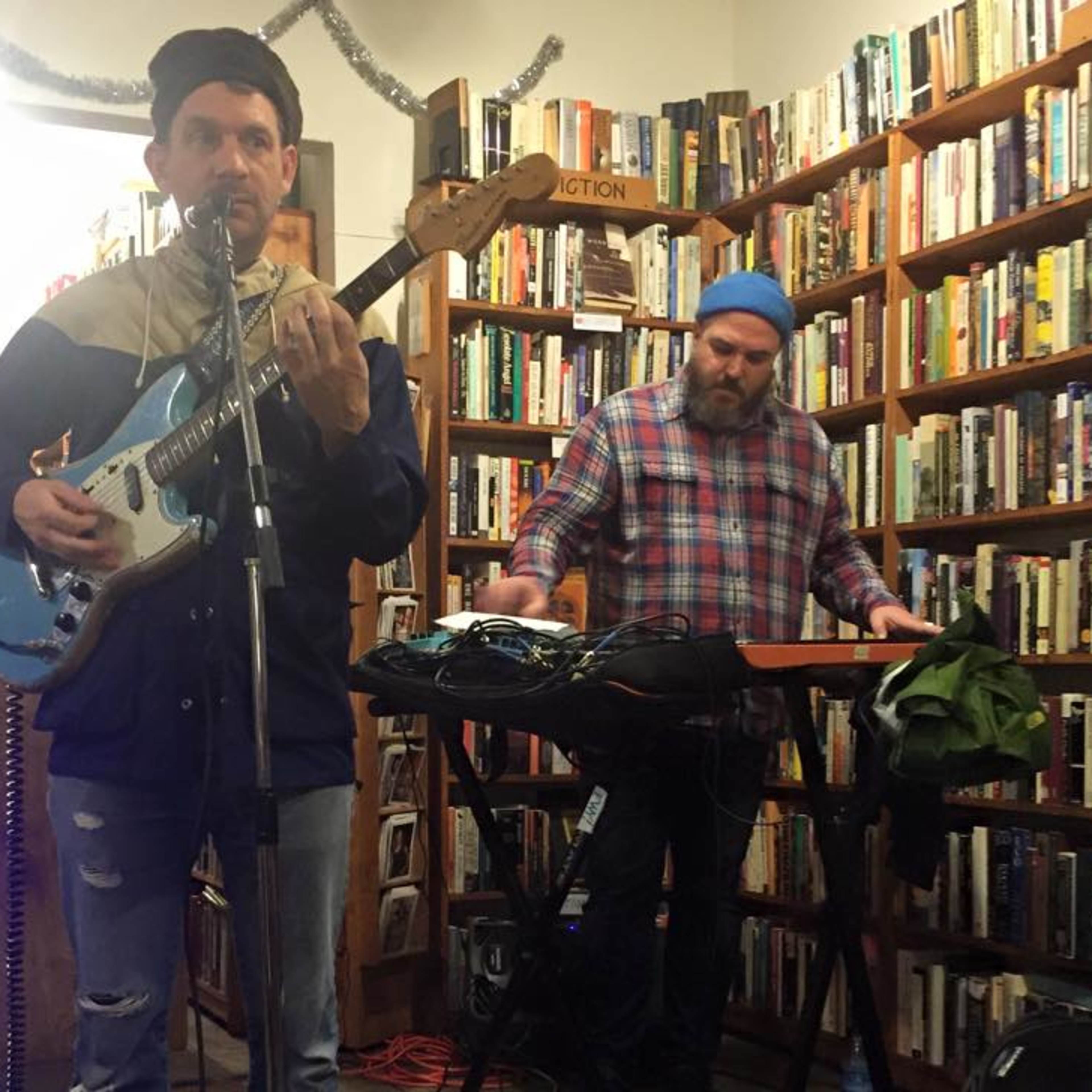 Two musicians perform in a cozy bookstore, one playing a guitar and the other using an electronic keyboard surrounded by shelves of books.